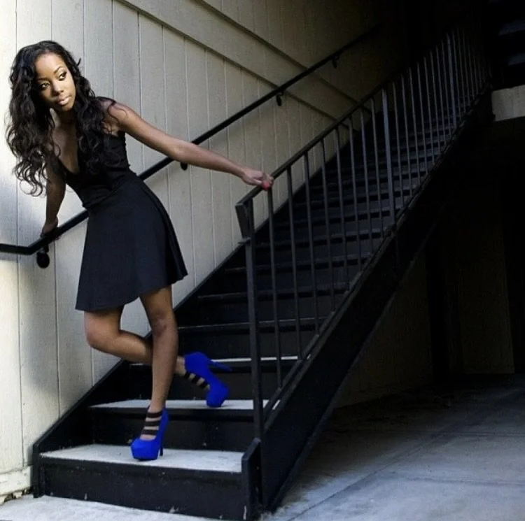 A young woman in a black dress and bright blue high heels stands on a staircase, holding the railing and posing with one leg bent. She has long, curly hair and is looking at the camera.