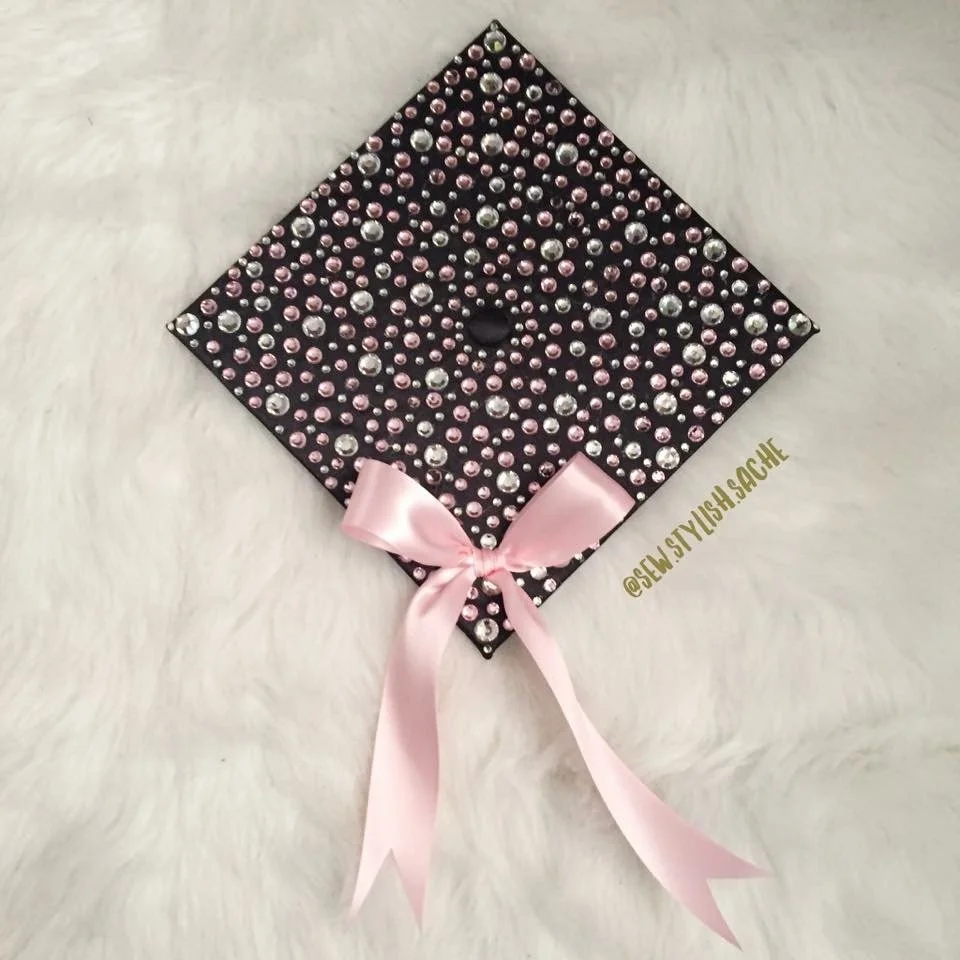Decorative graduation cap adorned with pink and silver rhinestones, featuring a pink satin bow, placed on a white furry surface.