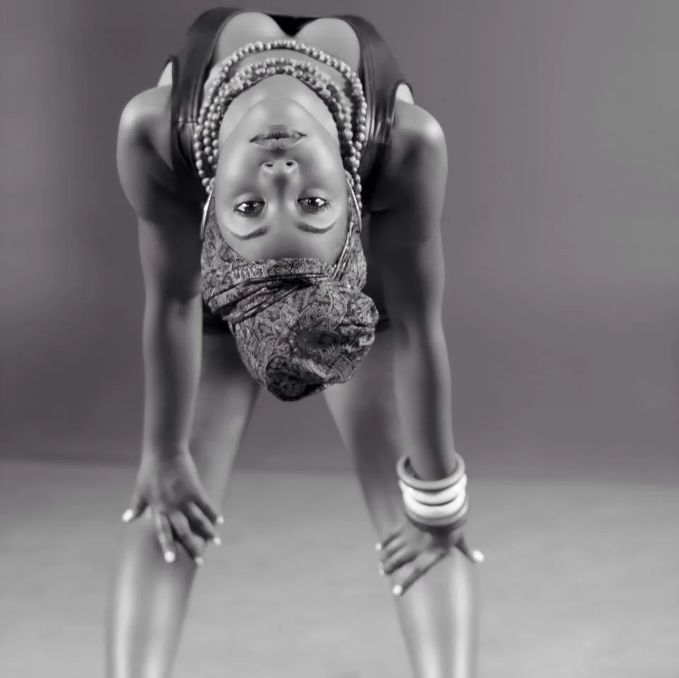 A woman in black and white is doing a yoga pose, bending forward with her hands on her knees, wearing a headwrap, beads, and bangles.