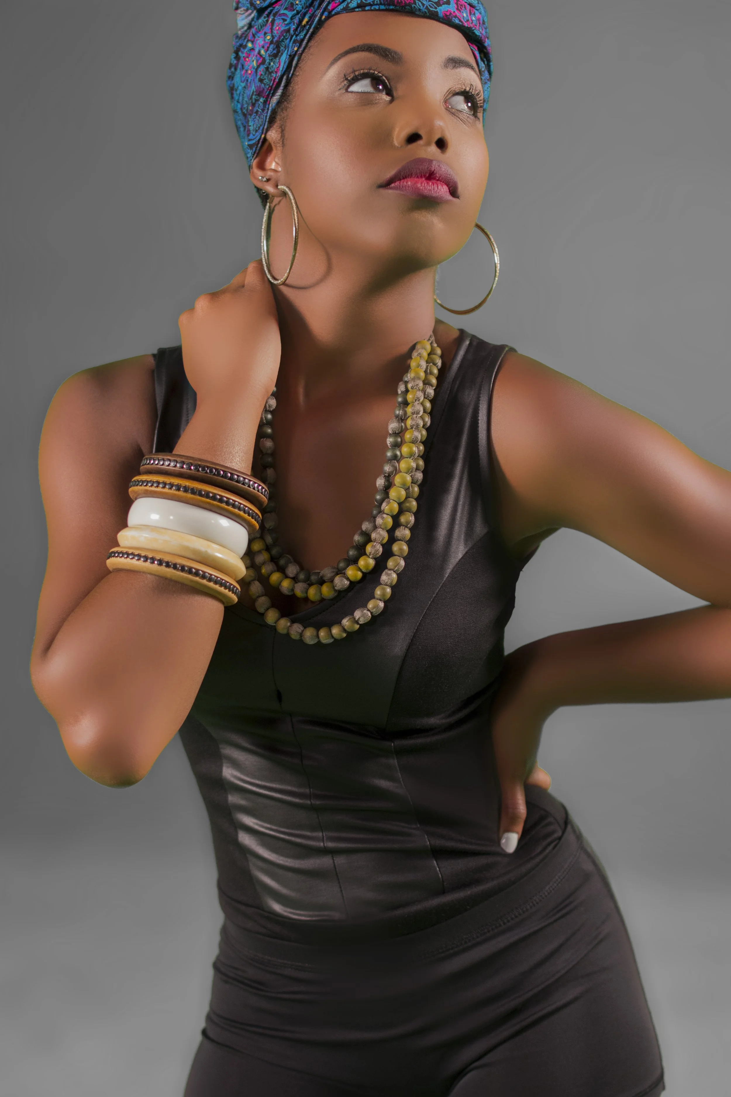 A woman wearing a colorful headscarf, large hoop earrings, and multiple bracelets, dressed in a black leather outfit, posing with her hand behind her neck against a gray background.