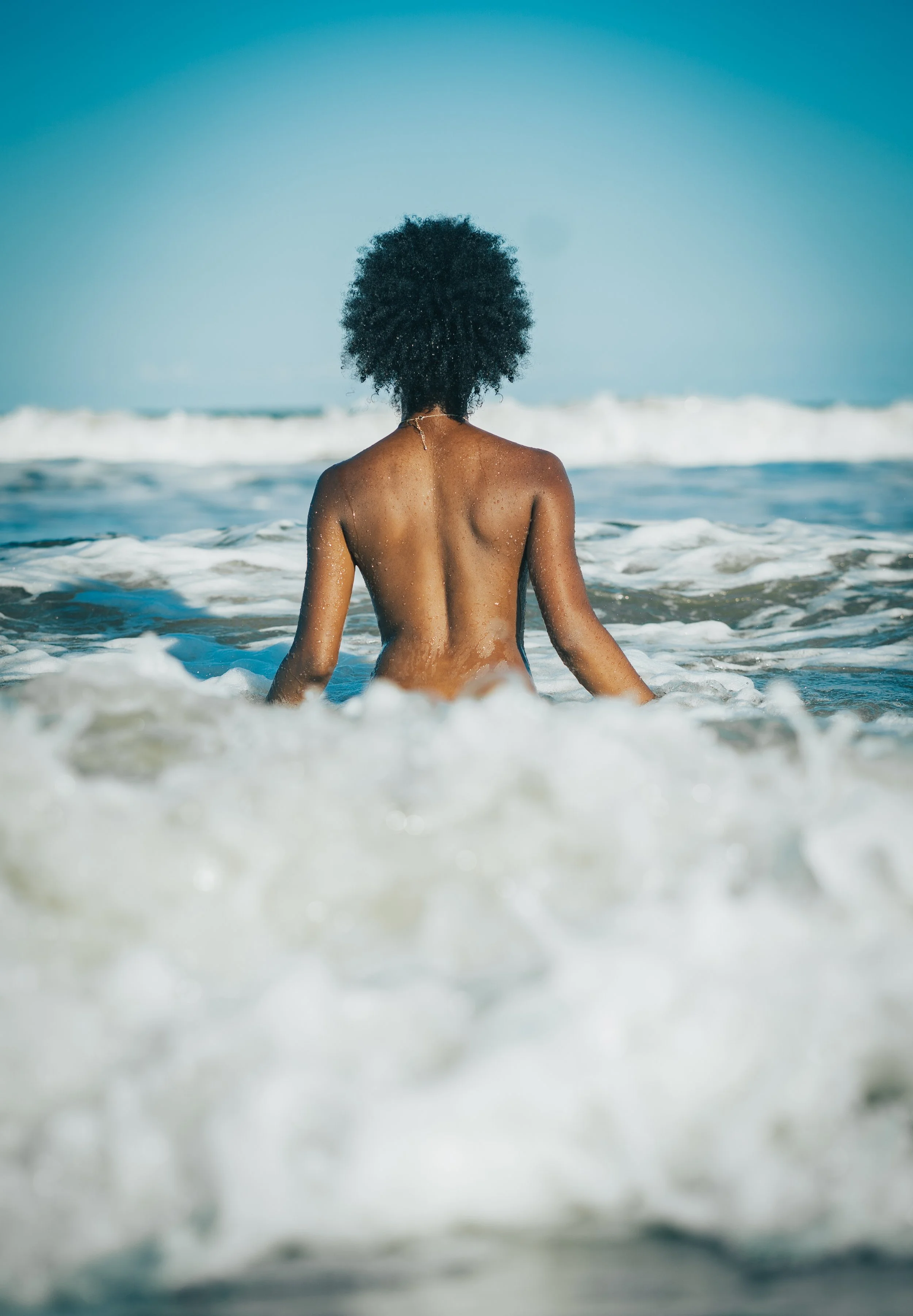 Back view of a woman with curly hair sitting in the ocean waves facing the horizon on a sunny day.