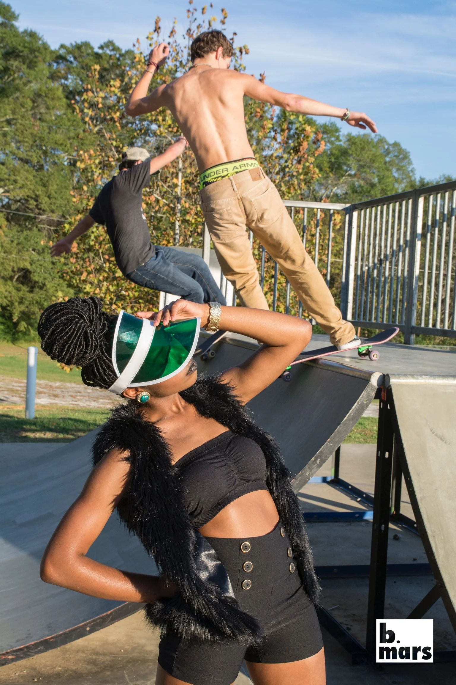 A woman with braided hair and large earrings wearing a black fur vest and high-waisted skirt posing at a skate park with skateboarding activity in the background. A shirtless man in tan pants and boxer briefs is skating on a ramp nearby, with trees a