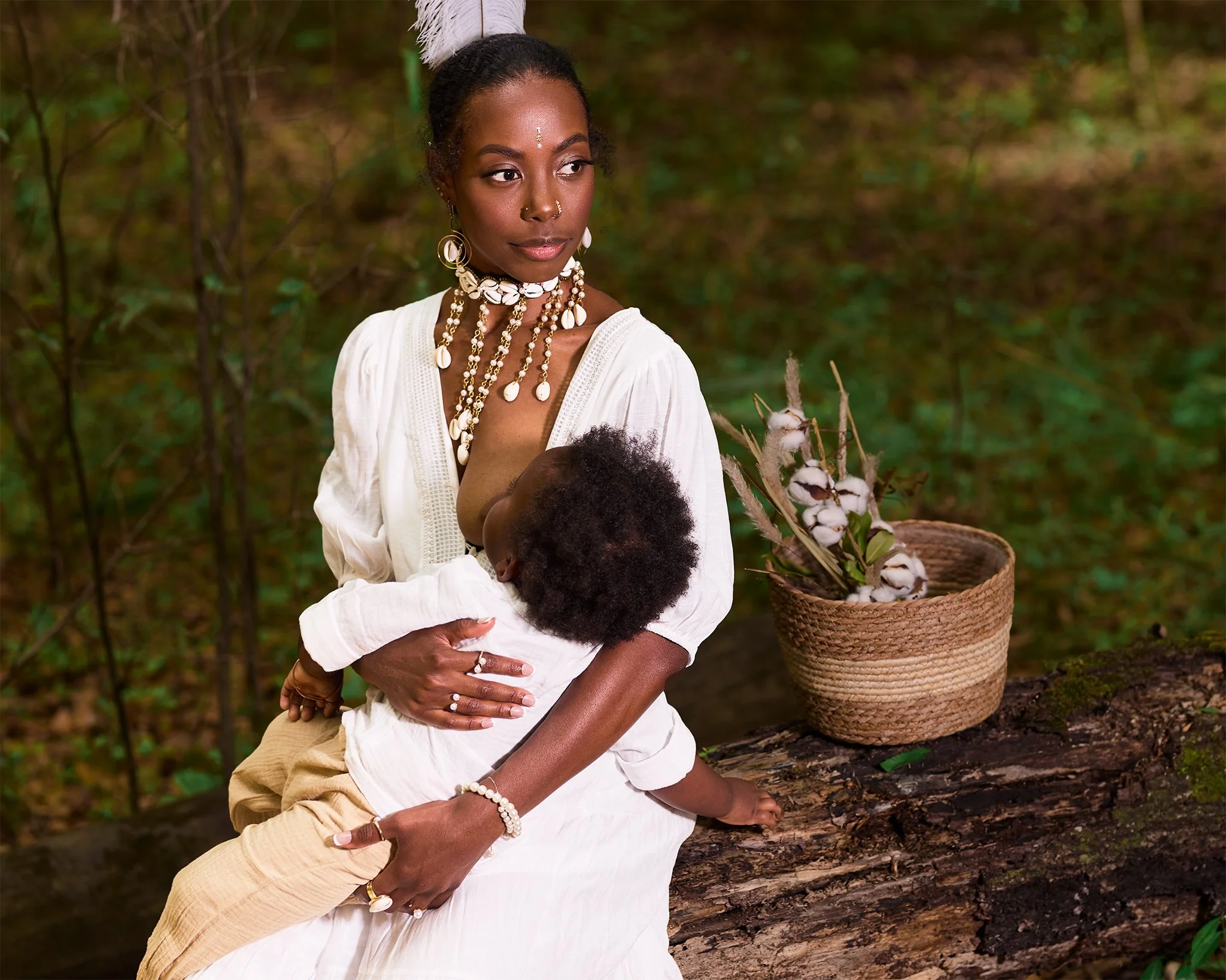 A woman and two children sit on a log in a forest, surrounded by greenery. The woman is wearing a white dress and ornate jewelry, holding a child who is breastfeeding. There is a basket of cotton and a woven basket nearby.