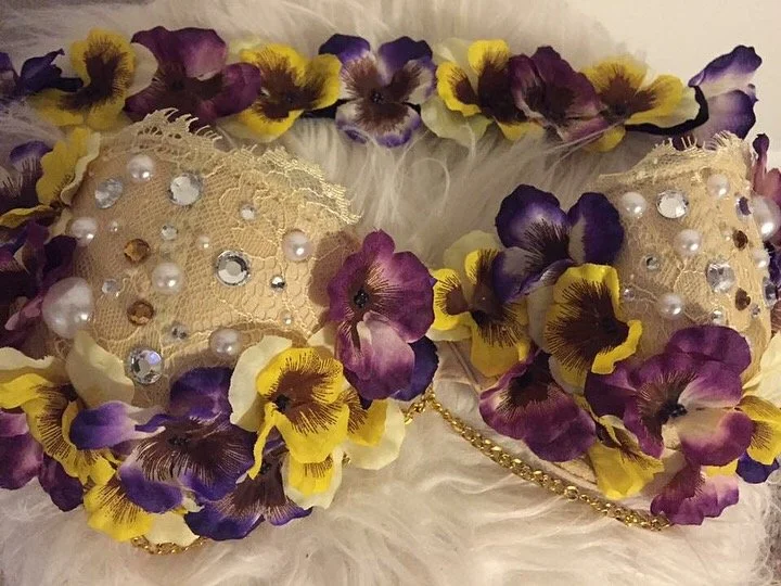 Decorative clutch purse adorned with purple, yellow, and cream artificial flowers, pearl beads, rhinestones, lace, and gold chain detail.
