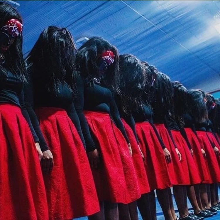 Group of women standing in a row, bowing their heads, wearing black tops and red skirts, with long dark hair and red face coverings, in a dimly lit indoor setting.