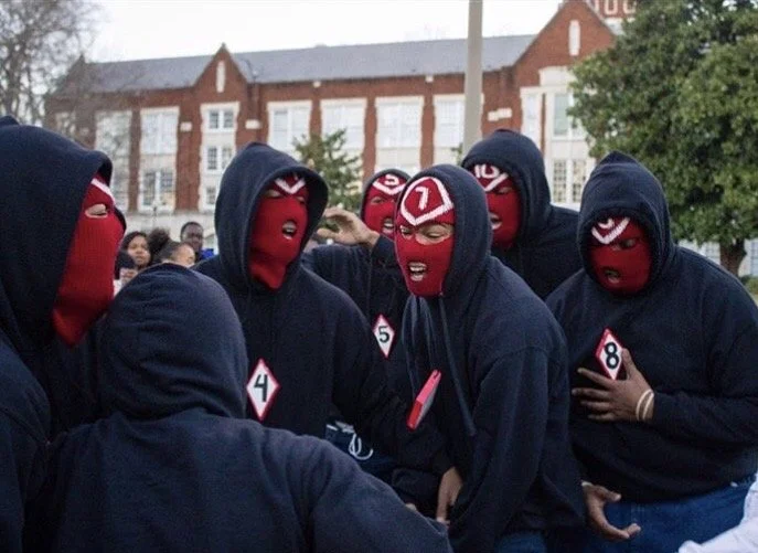 Group of people wearing red masks with number tags, gathered outdoors, wearing dark hoodies, possibly in a protest or demonstration.