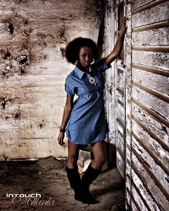 Young woman with curly hair wearing a blue denim dress and black boots, standing in a rustic, weathered room with wooden walls and dirt floor, posing with one arm resting on a slatted wall.