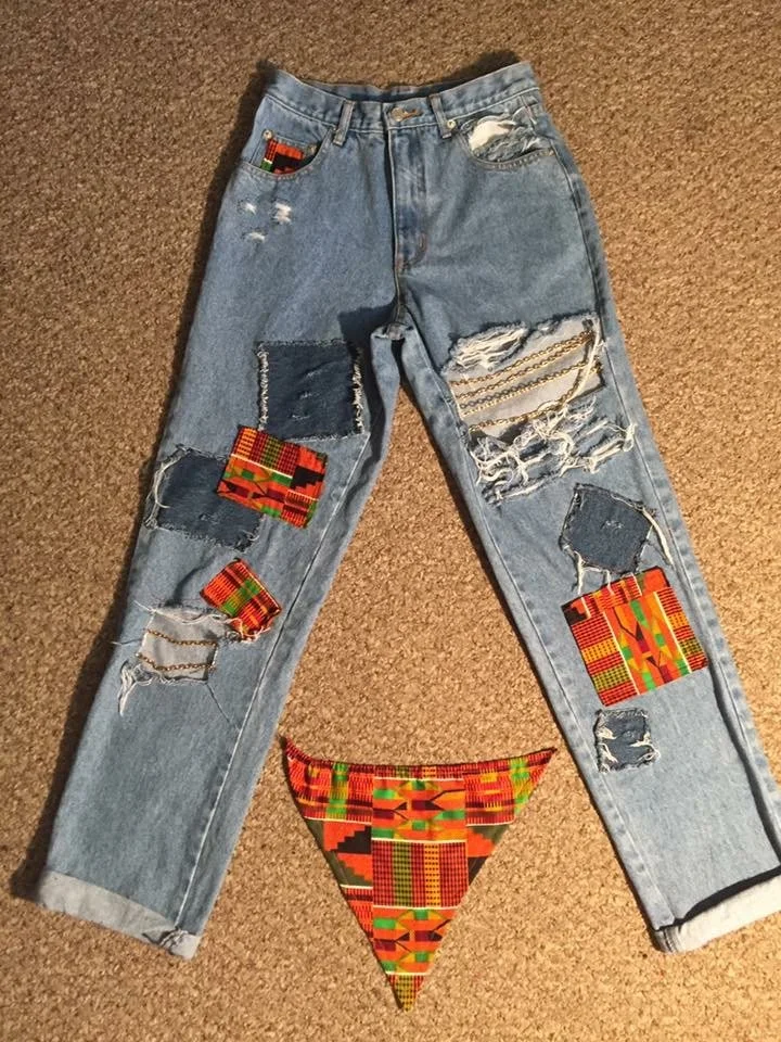 A pair of light blue, distressed jeans with various colored African fabric patches sewn onto them, lying flat on a beige carpet. There is also an African fabric face mask with similar colorful patterns placed below the jeans.