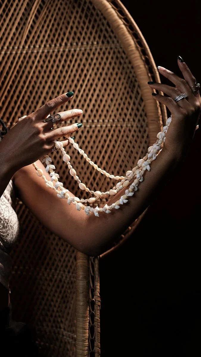 Person holding a pearl necklace in front of a wicker mirror, with a dark background.