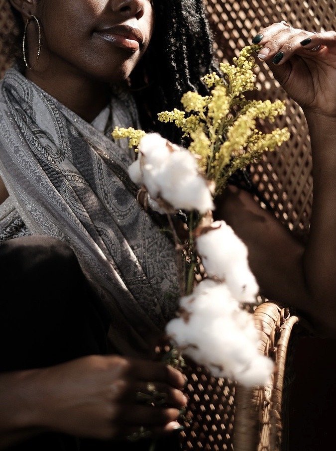 A woman holding a bunch of yellow flowers and cotton stems, with a woven chair and a wooden lattice background.
