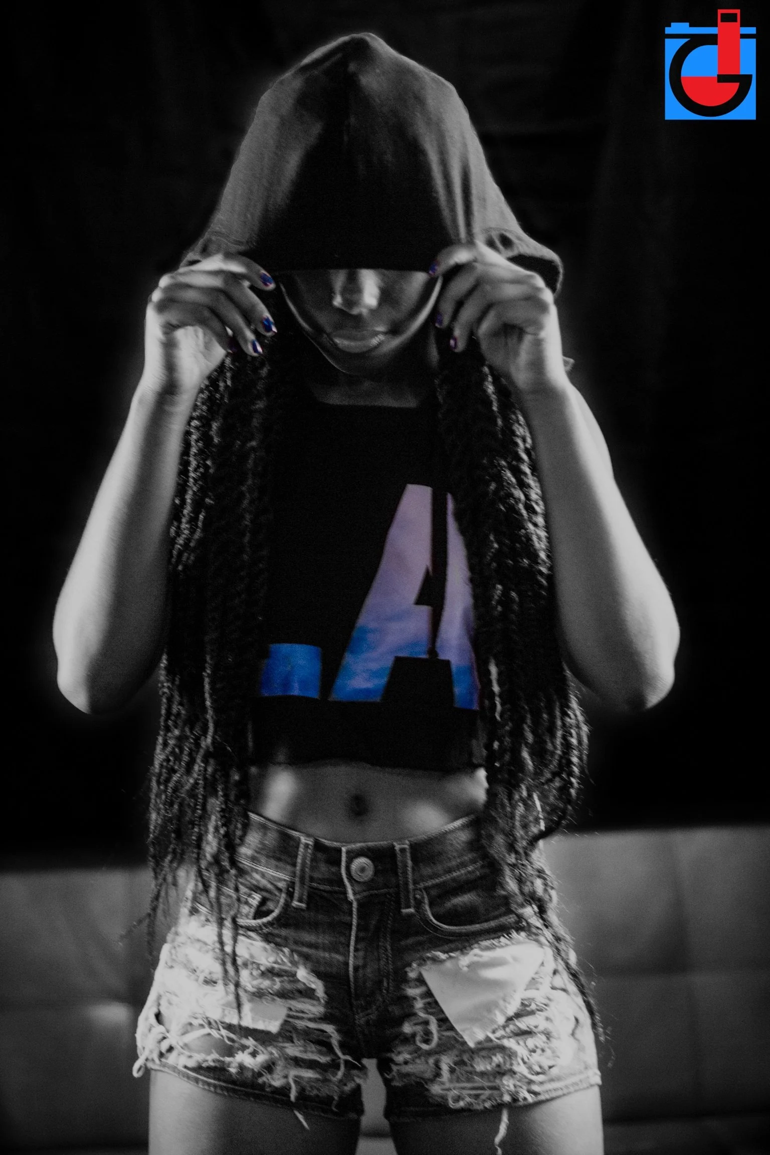 A person wearing a hooded sweatshirt with the hood pulled over their head and face, holding onto the hood with both hands, and wearing distressed denim shorts and a graphic T-shirt.