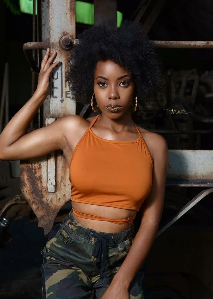 A young woman with natural hair wearing an orange crop top and camouflage pants, posing in front of a rusty industrial background.