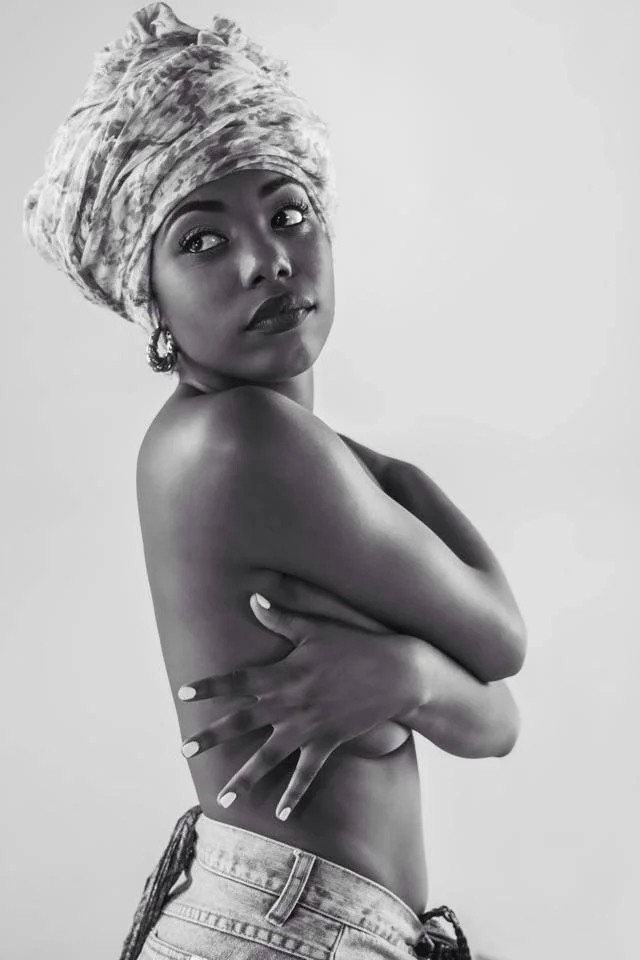 A black and white photo of a woman with a head wrap, earrings, and painted nails, posing topless with her arms crossed over her chest.
