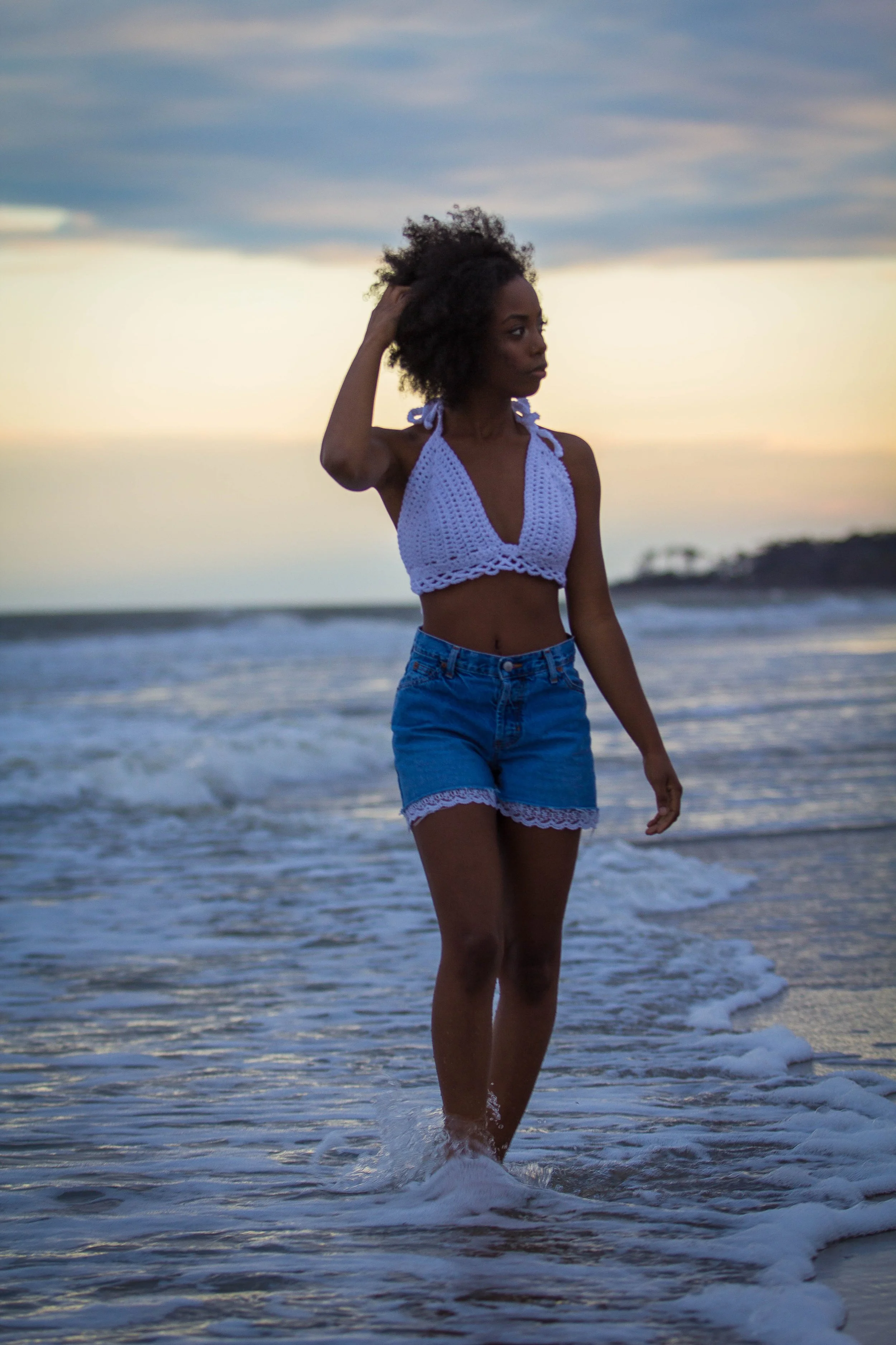 A woman standing in the ocean at sunset, wearing a white crochet top and denim shorts, with curly hair and a hand touching her head.