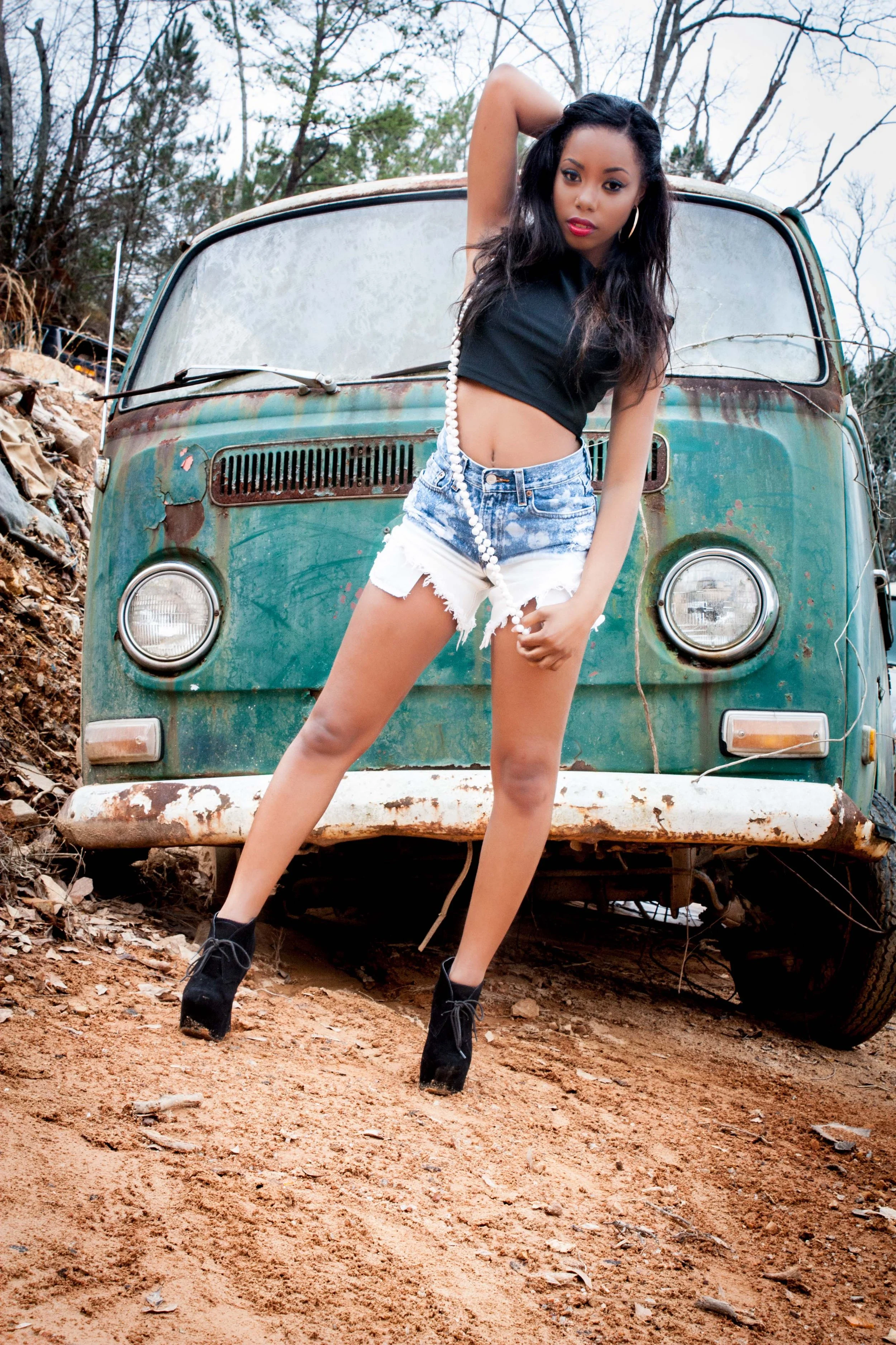 A young woman with dark hair, wearing a black crop top, distressed denim shorts, and black high heels, stands in front of an old, weathered Volkswagen van outdoors on a dirt surface with trees in the background.