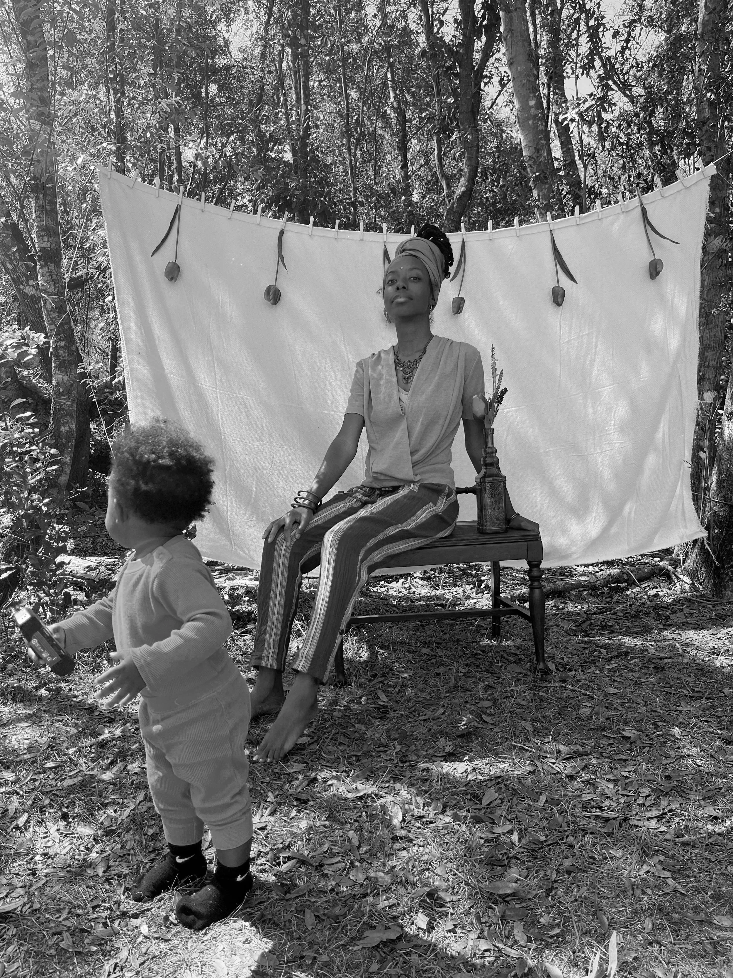 Black-and-white photo of a woman sitting on a wooden stool behind a white sheet in a wooded area, with a child in the foreground holding a toy or device, and trees in the background.