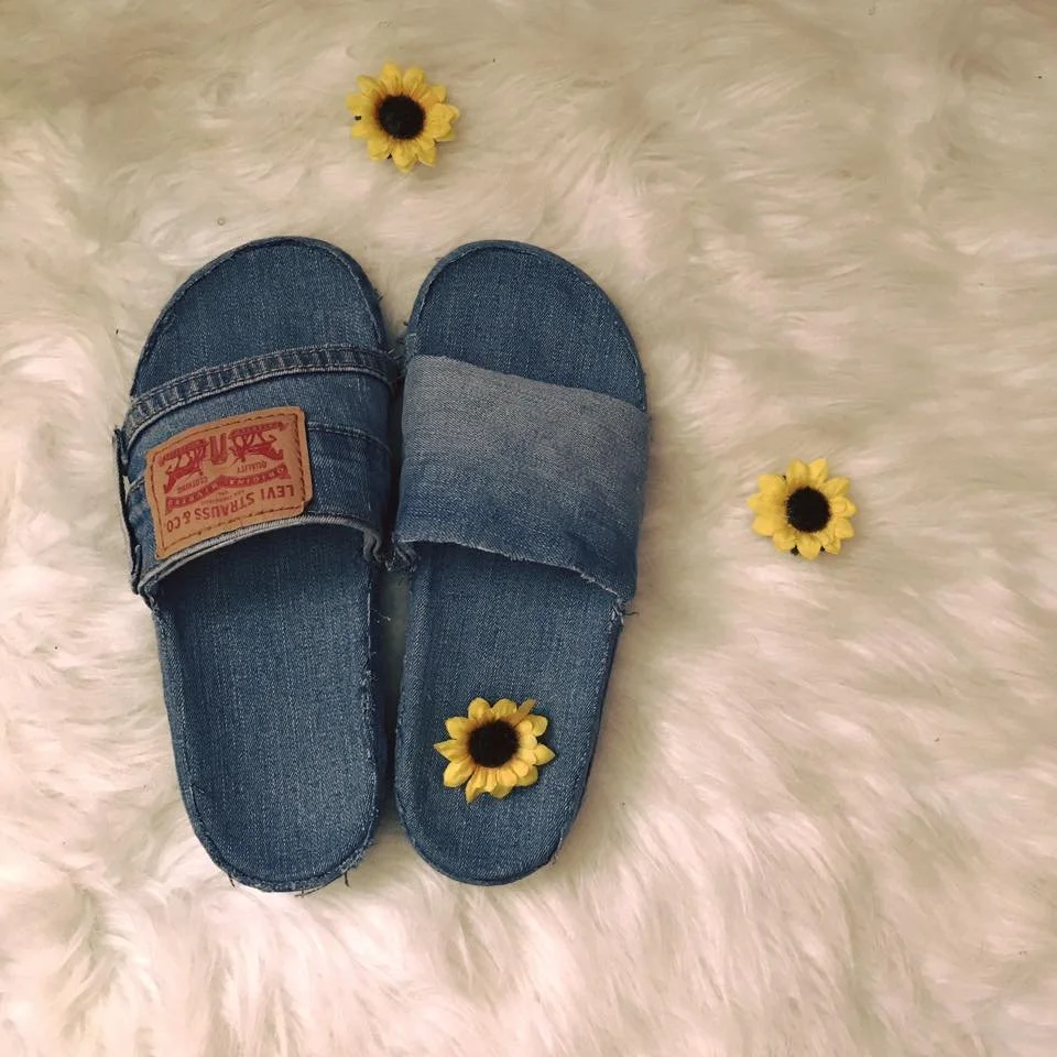 Pair of denim slippers with Velcro straps on a white fluffy surface, surrounded by three small yellow flowers with black centers.