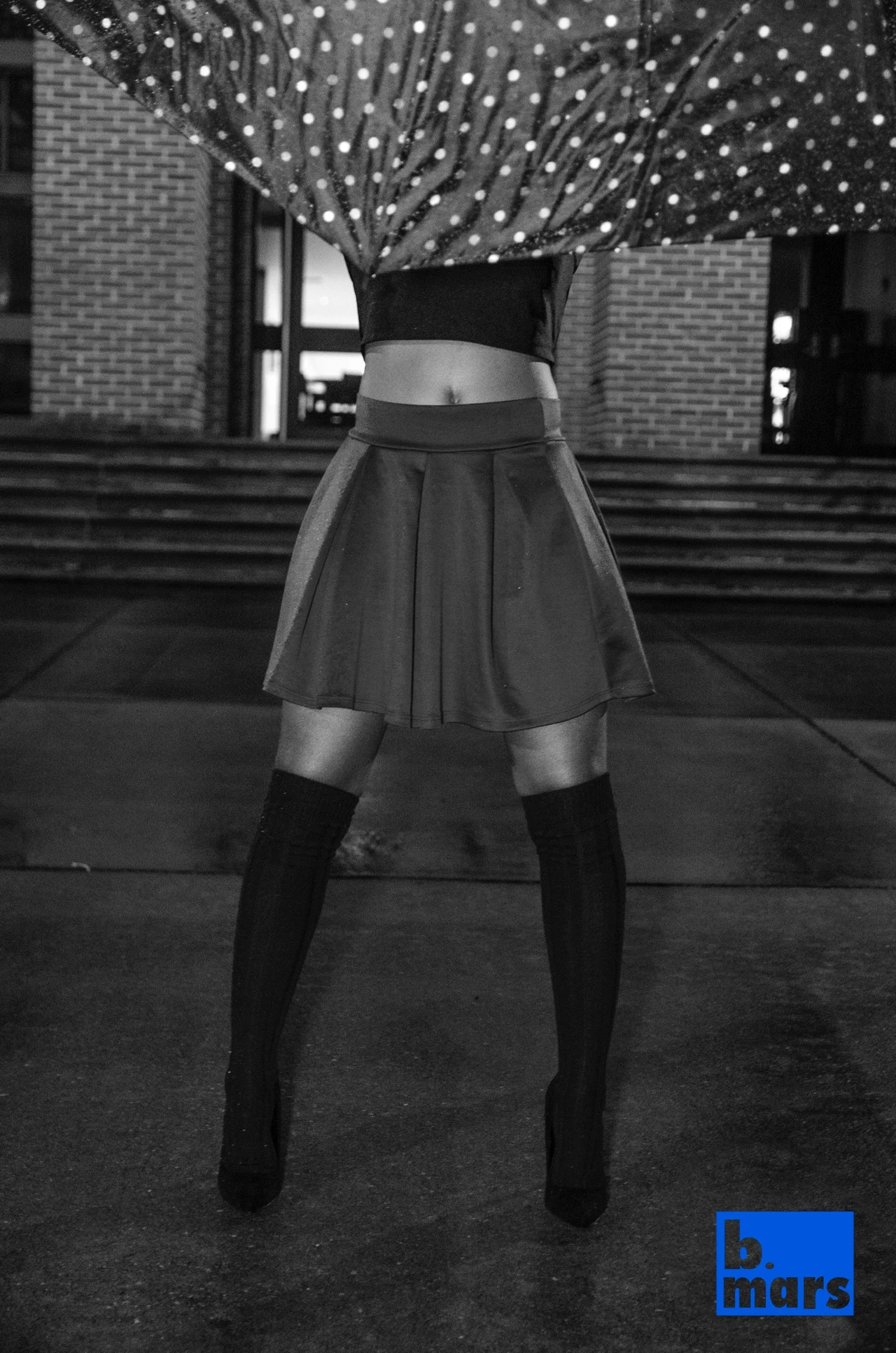 A person standing outdoors holding an umbrella with their face obscured. The person is wearing a skirt and thigh-high socks. The background includes brick walls and steps, and a blue rectangular logo with the words 'b. mars' in the bottom right corne