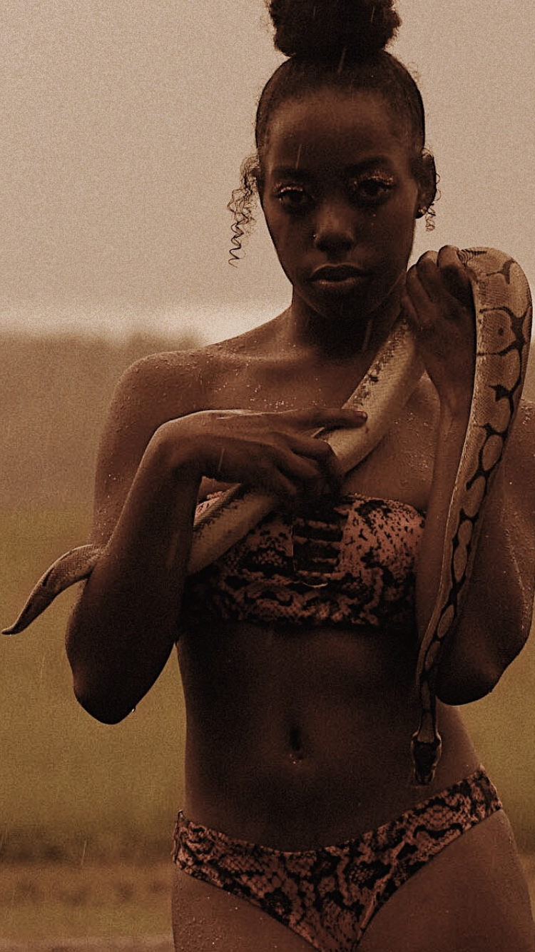 A woman with curly hair in a high bun, wearing matching animal print bikini, holding a snake around her shoulders outdoors in a natural setting.