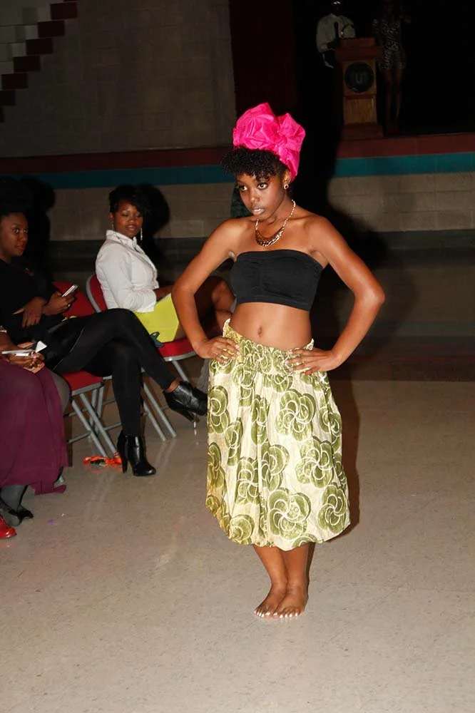 Young woman performing a traditional dance, dressed in a black crop top, gold patterned skirt, and wearing a large pink head wrap, with seated women in the background watching.