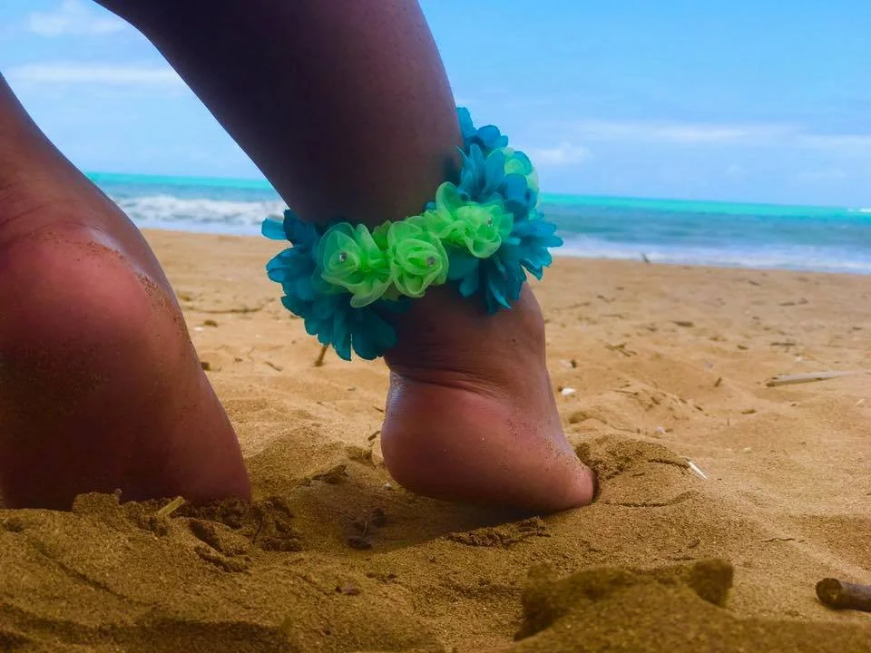 Close-up of a person's feet and legs on a sandy beach, with an ankle decoration made of blue and green flowers, and the ocean in the background.