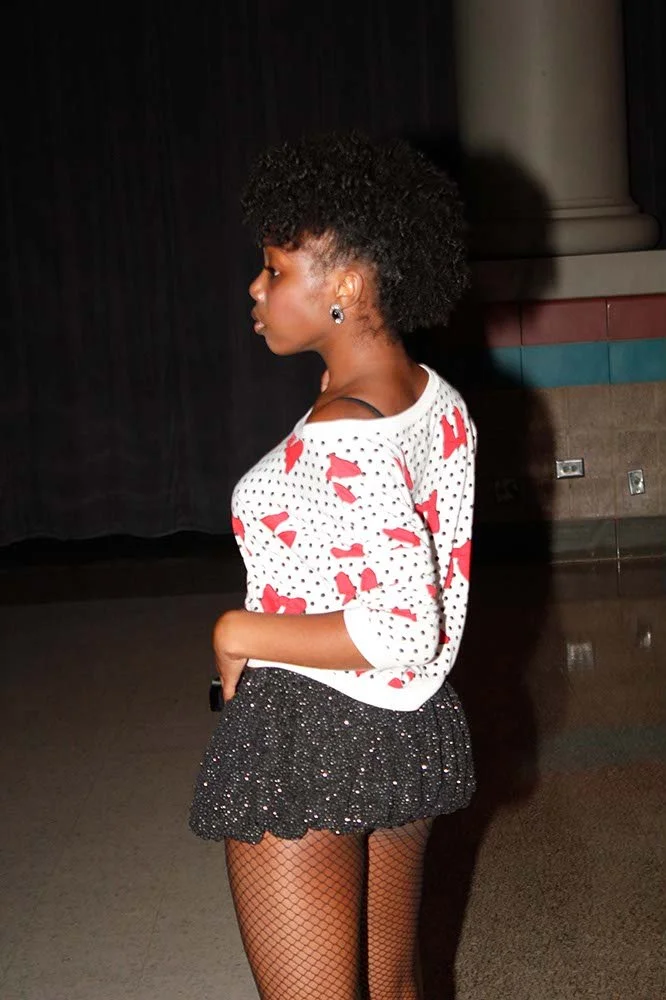 Side view of a young woman with curly black hair wearing a white sweater with red pattern, a black sparkly skirt, and fishnet stockings, standing indoors with dark background and columns.