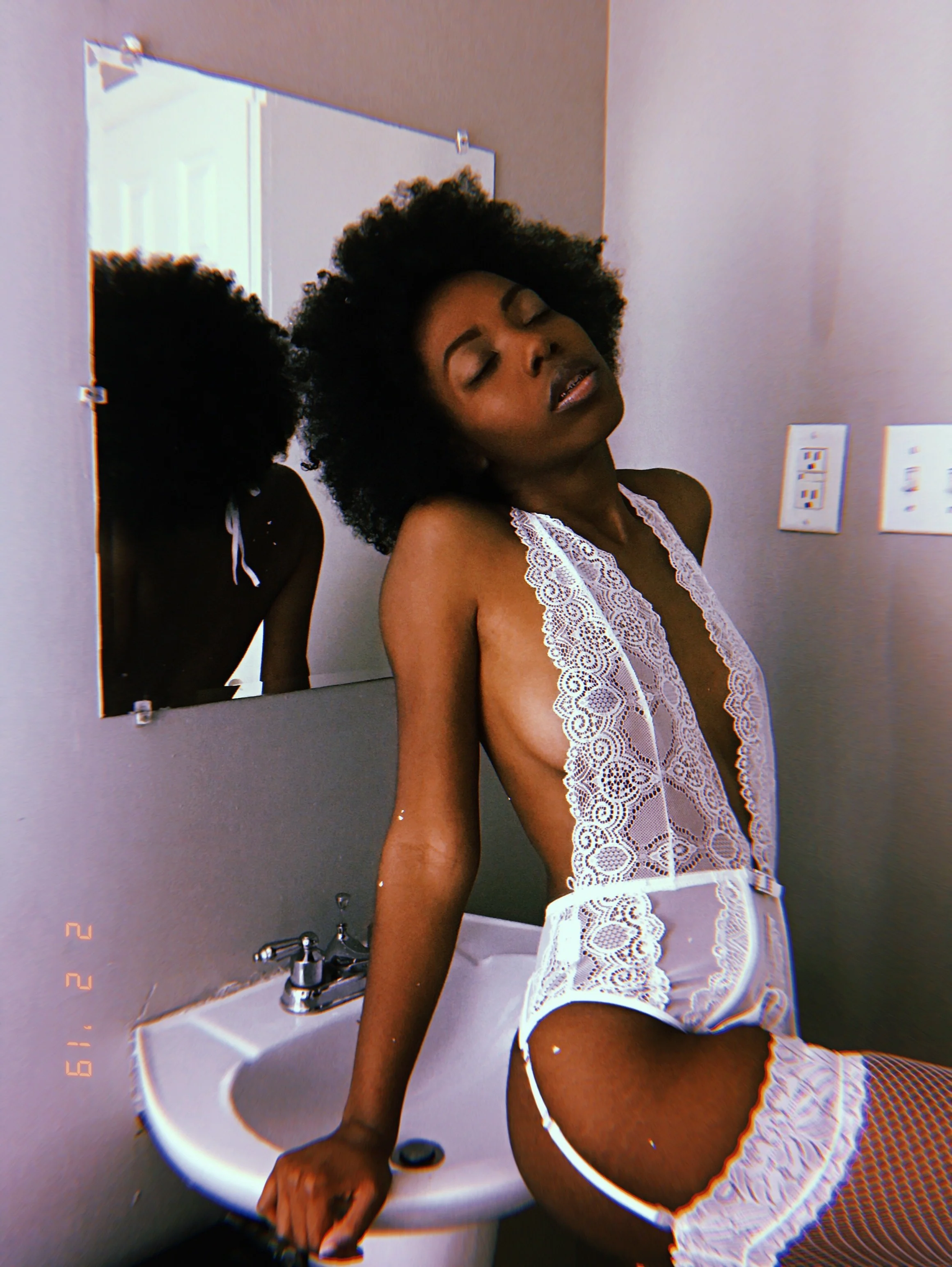 A woman with natural curly hair in a bathroom, wearing white lace lingerie, with her eyes closed, standing next to a bathroom sink and mirror.