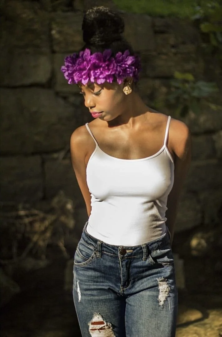 A young woman with dark hair in a high bun adorned with a purple flower crown, wearing gold earrings, a white spaghetti strap top, and distressed jeans, standing outdoors with a stone wall background, looking downward.