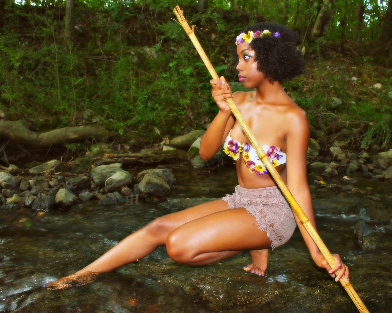 A woman with curly hair, wearing a flower crown and a floral crop top, sitting in a shallow stream holding a bamboo stick in a lush green forest.