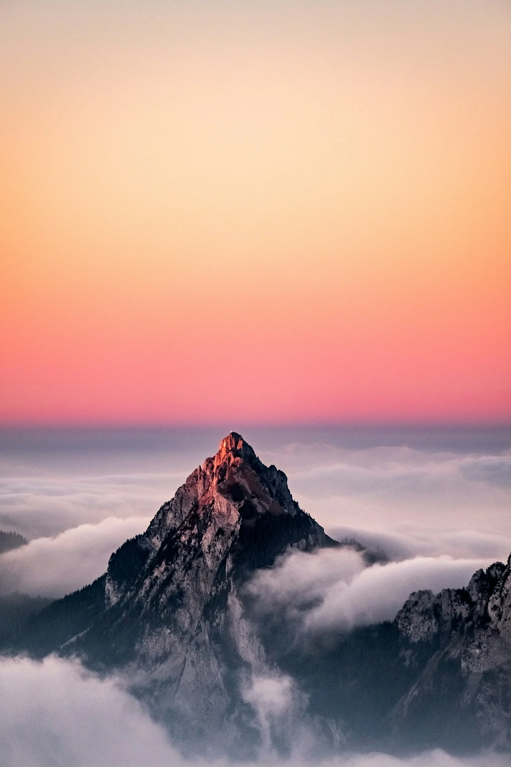 A tall mountain peak rising above the clouds during sunset or sunrise, with pink and orange hues in the sky.