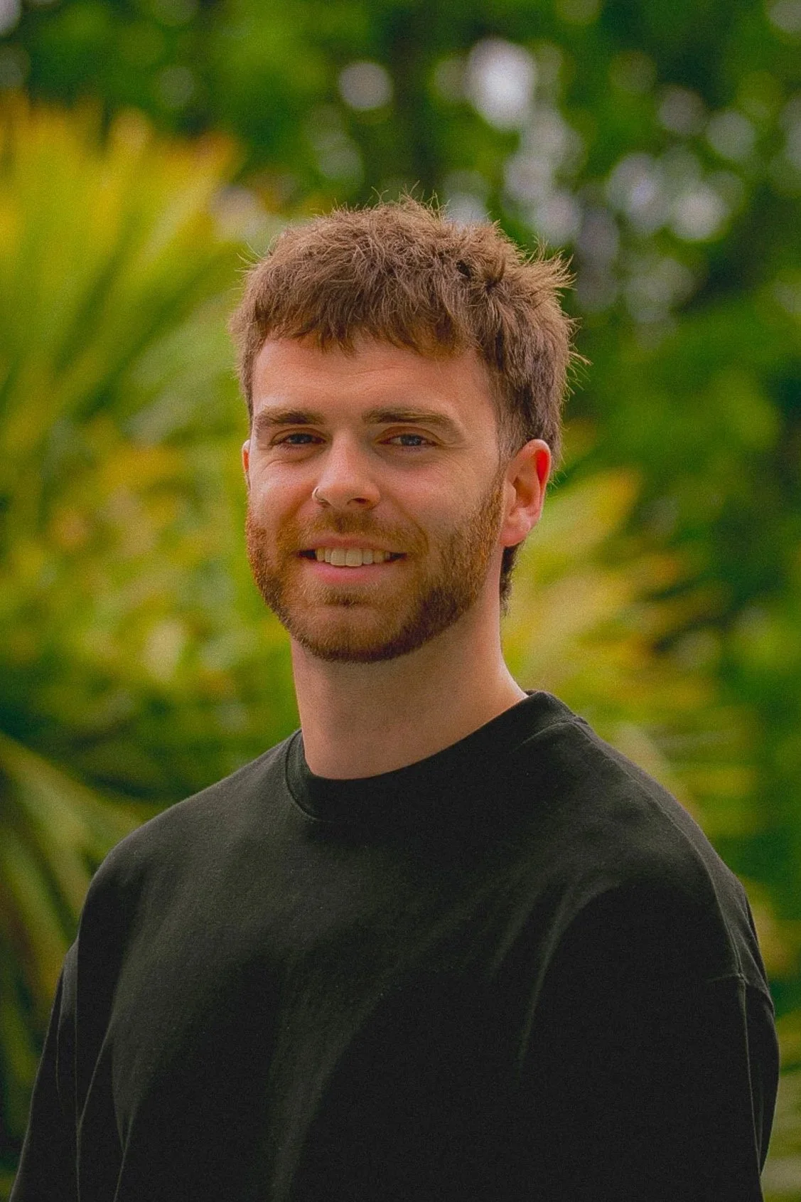 A young man with brown hair and a beard smiling outdoors with a background of green foliage.