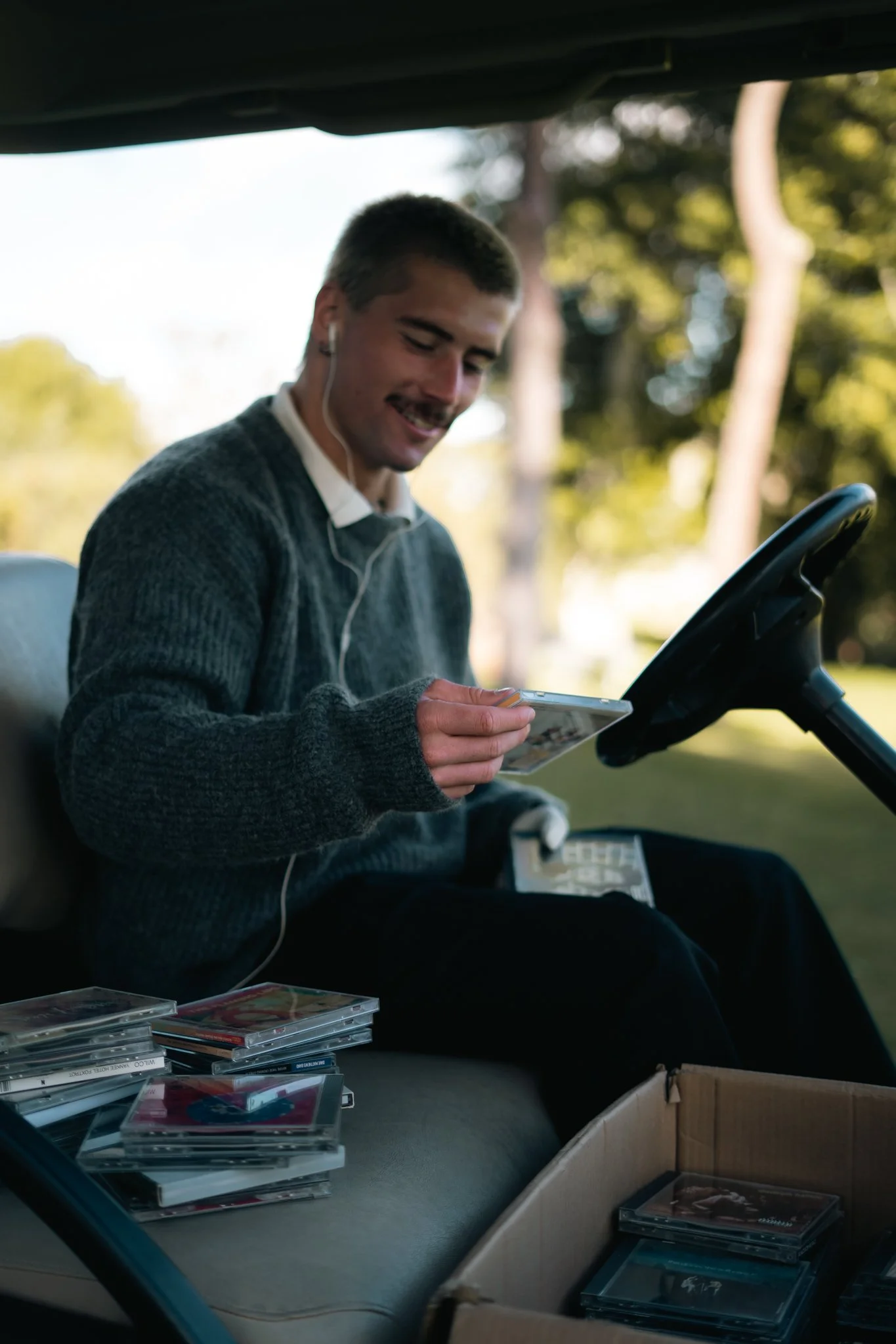 A young man with short dark hair, wearing a gray sweater and white earphones, smiling while looking at his phone, sitting inside a golf cart. The cart's dashboard and a box of CDs or DVDs are visible, with more CDs or DVDs in plastic cases on the seat. Outdoors with a backdrop of trees and sunlight.