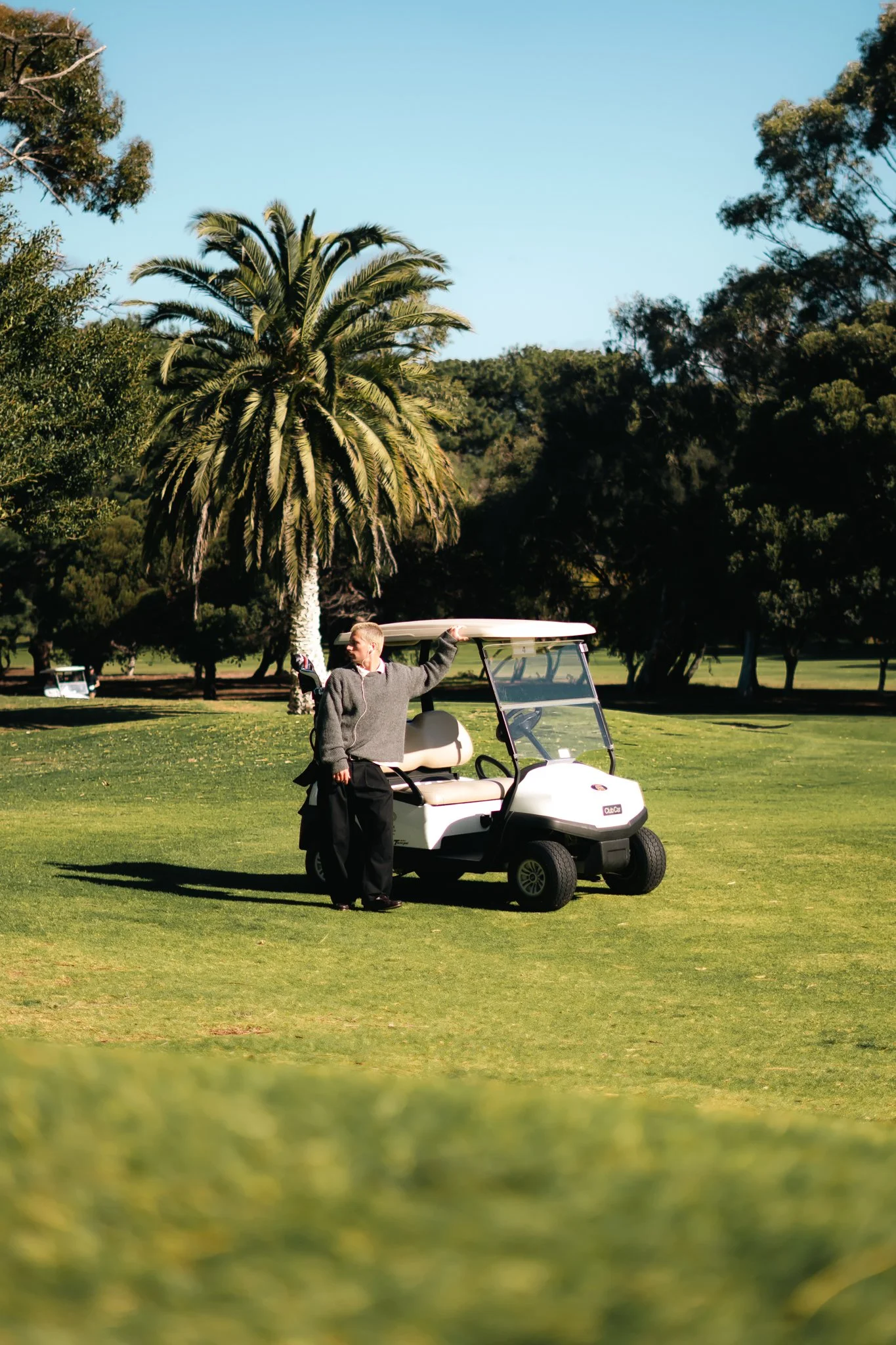 A person standing next to a white golf cart on a golf course, with palm trees and other trees in the background on a sunny day.
