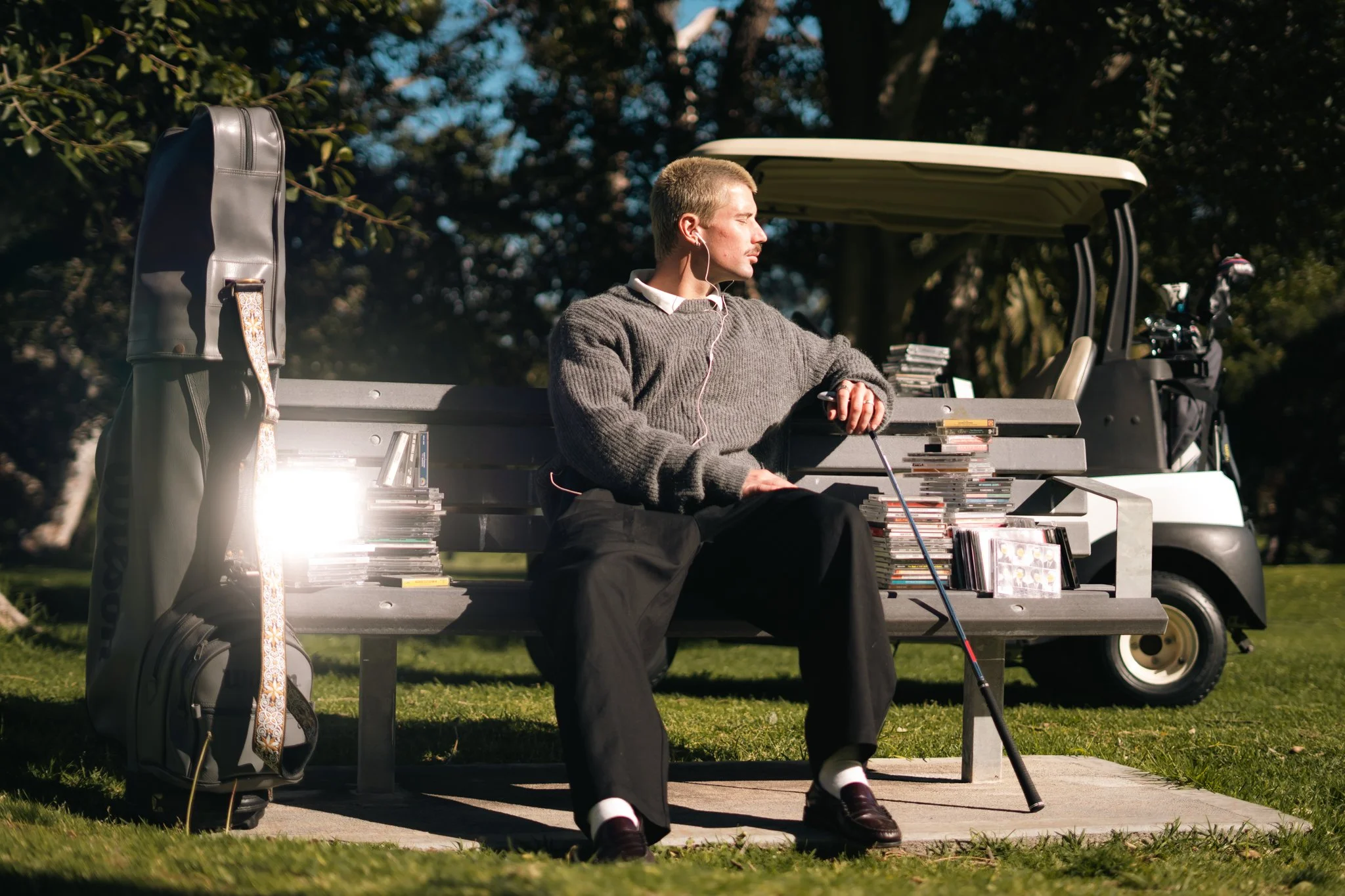 A young man sitting on a park bench, wearing a sweater, listening to music through earphones, with a golf bag and golf clubs nearby, and a golf cart in the background.