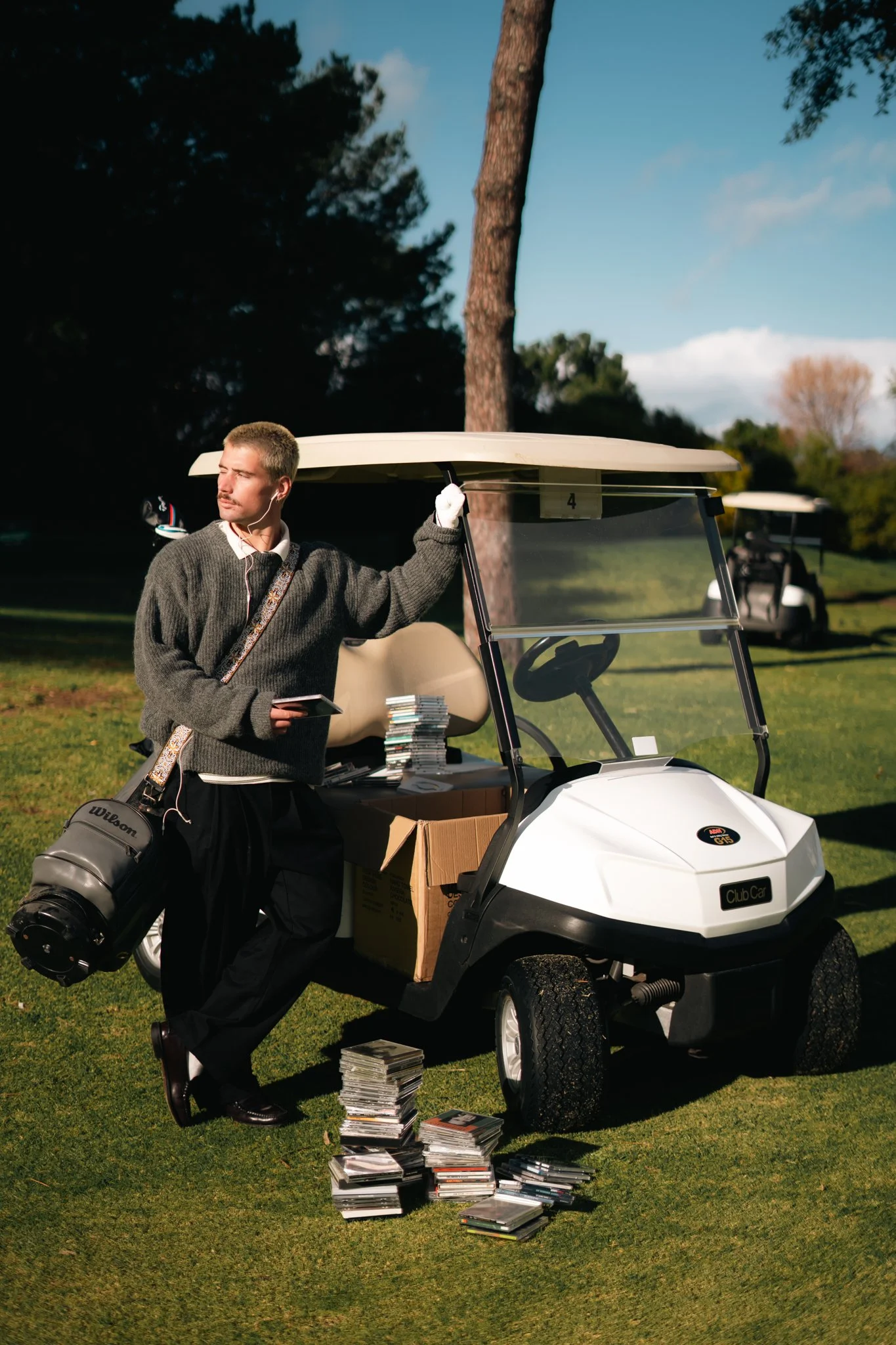 A man with short hair and a mustache wearing a gray sweater, white shirt, and white gloves, is standing beside a white golf cart on a golf course. He is holding the golf cart roof with one hand, and has earbuds in his ears. There are stacks of magazines or papers on the ground next to the cart, with more stacked on the cart's seat. The golf course has green grass, trees, and a blue sky with some clouds in the background.