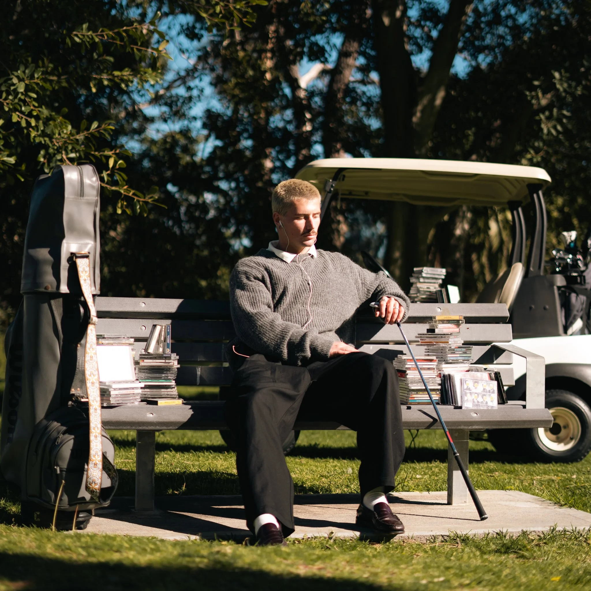 A man wearing a gray sweater and black pants is sitting on a bench outdoors, surrounded by trees. He appears to be relaxing, with a golf club resting on his lap, and a golf bag and a stack of CDs or DVDs nearby. The scene is set on a sunny day.