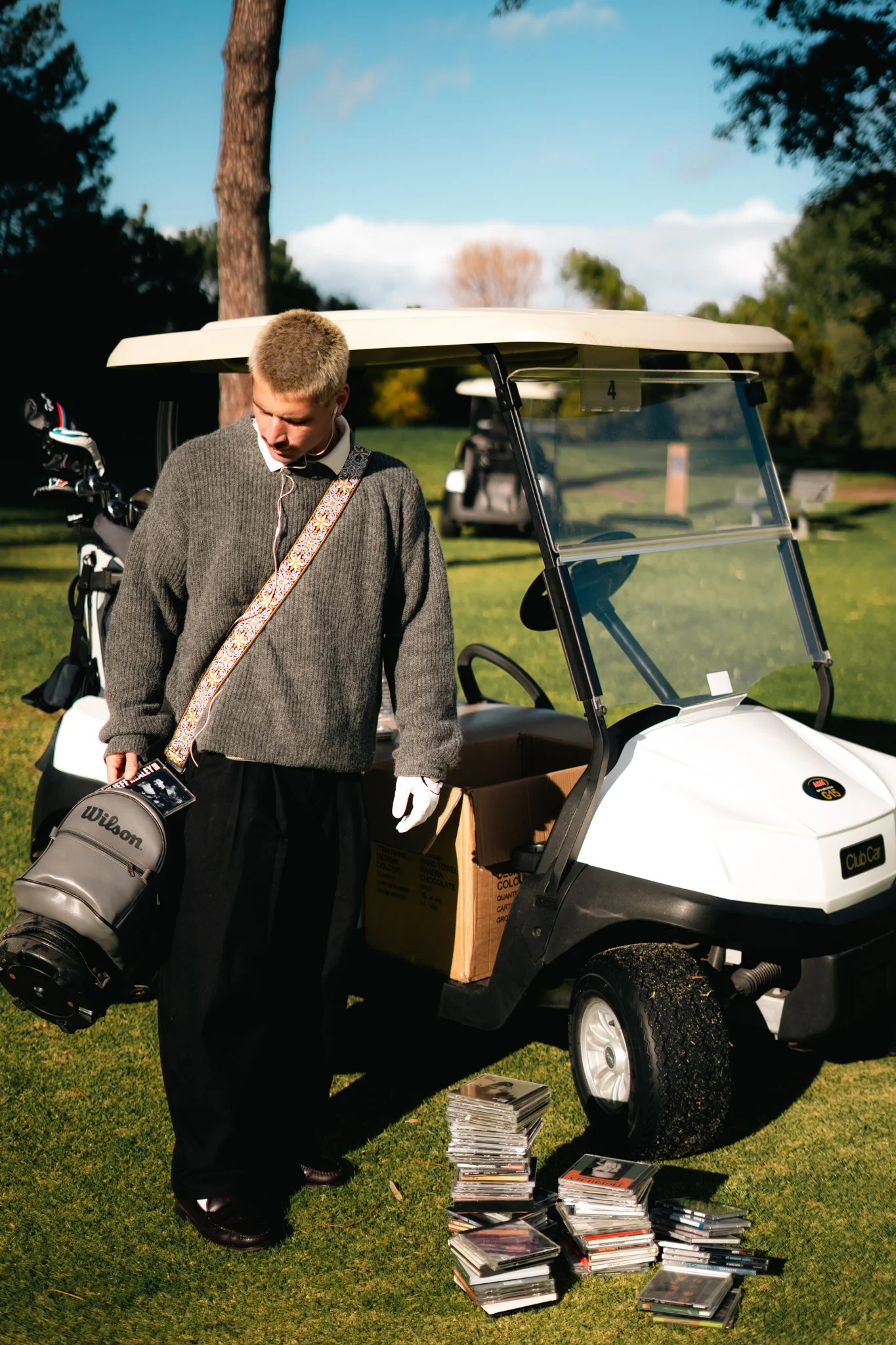 A young man wearing a gray sweater and dark pants taps a magazine on the ground with several stacks of magazines and books in front of him. He stands next to a white golf cart with a visor and a cart bag of golf clubs attached. The background shows green grass, trees, and a blue sky with some clouds.