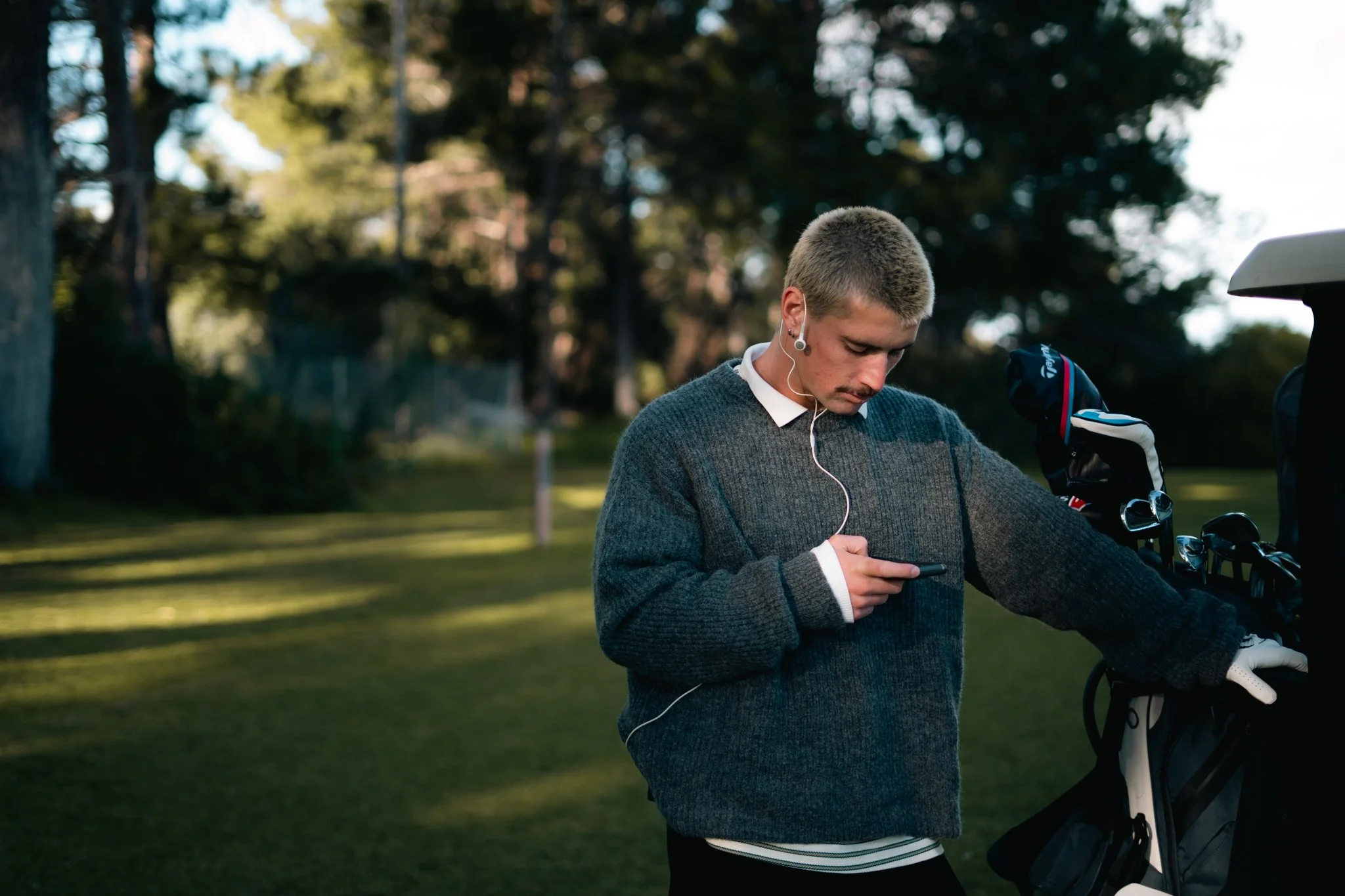 A young man with short blond hair, wearing a gray sweater over a white shirt, stands outdoors with a golf club and glove, looking at his smartphone while listening to earphones. The background shows trees and a grassy area, suggesting he is on a golf course.