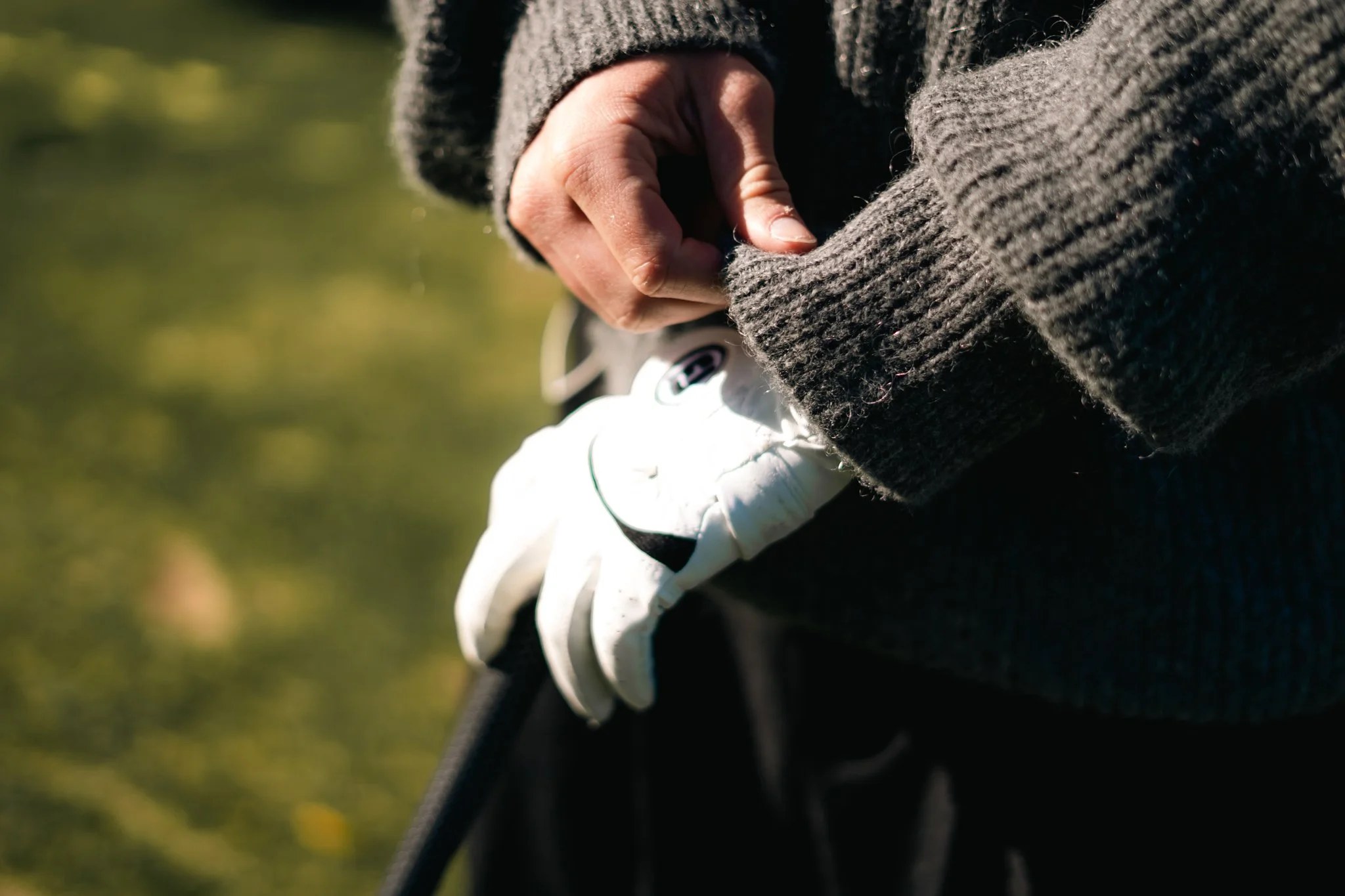 A person tying the shoelaces of their white athletic shoes while wearing black pants and a gray sweater. The background is blurred but suggests an outdoor setting with greenery.