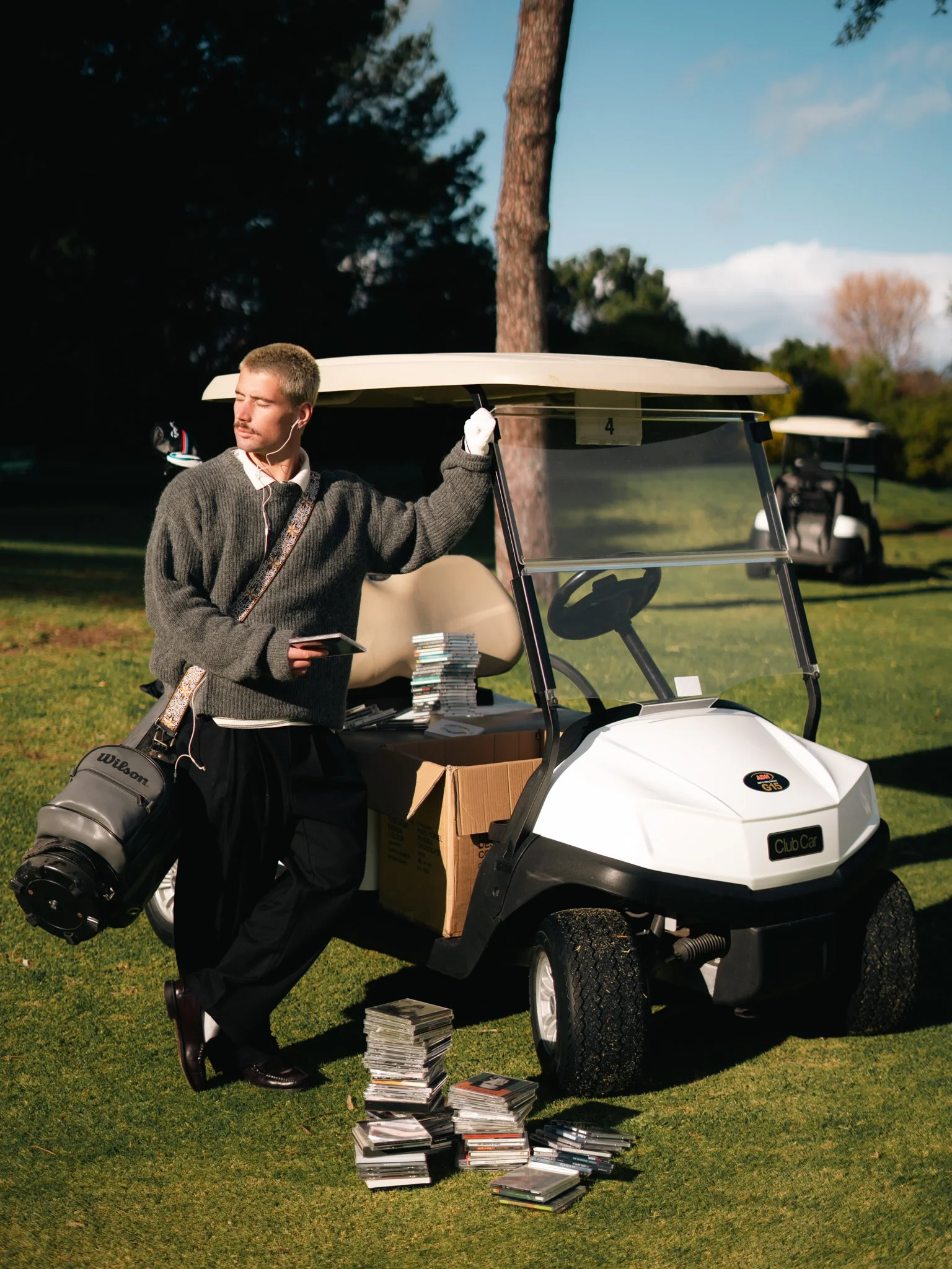 A man with a mustache in a gray sweater and black pants stands next to a white golf cart filled with stacks of magazines, holding a smartphone in his right hand and a golf glove in his left. There is a golf bag attached to the cart with a golf club sticking out. The scene is outdoors on a grassy area with tall trees and a blue sky in the background. Another golf cart is visible in the distance.