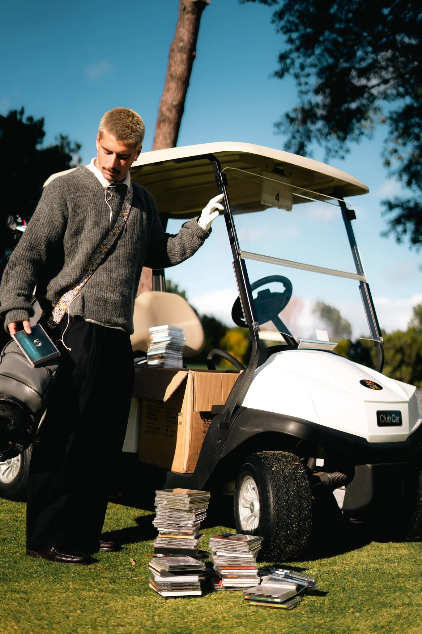 A man arranging a large collection of CDs on the ground next to a golf cart with a stack of CDs on top, outdoors on a sunny day with trees in the background.