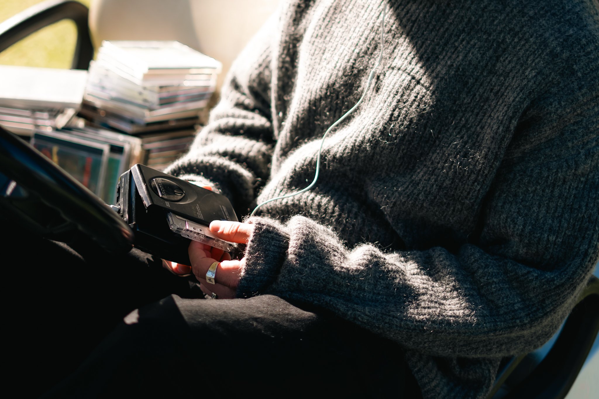 Person wearing a gray knitted sweater using a smartphone with a small external microphone attached, sitting near a stack of CDs or DVDs.