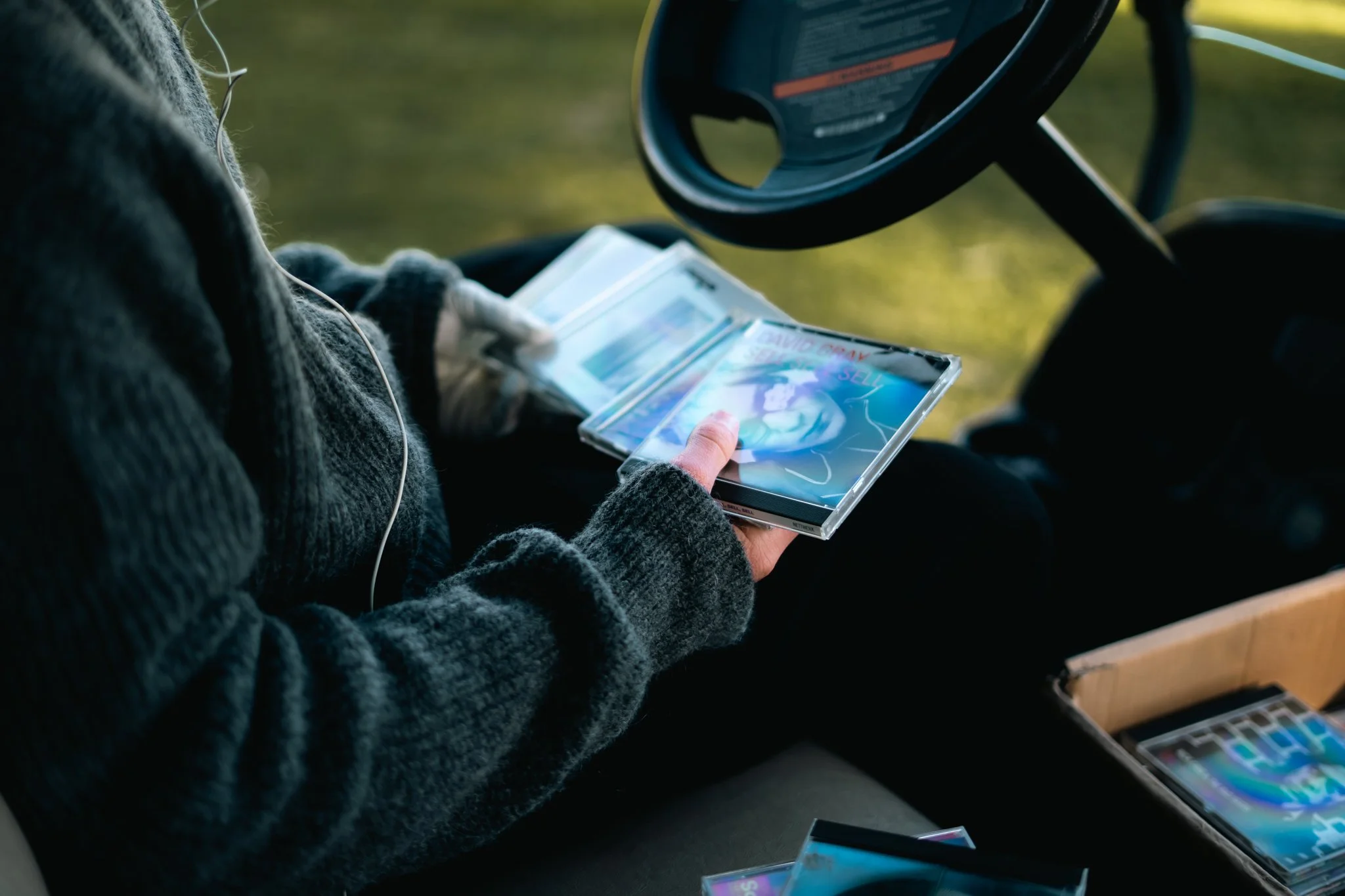Person wearing a dark grey sweater with white earphones, holding a CD case with colorful artwork, sitting at a table with more CD cases and a color palette in front.