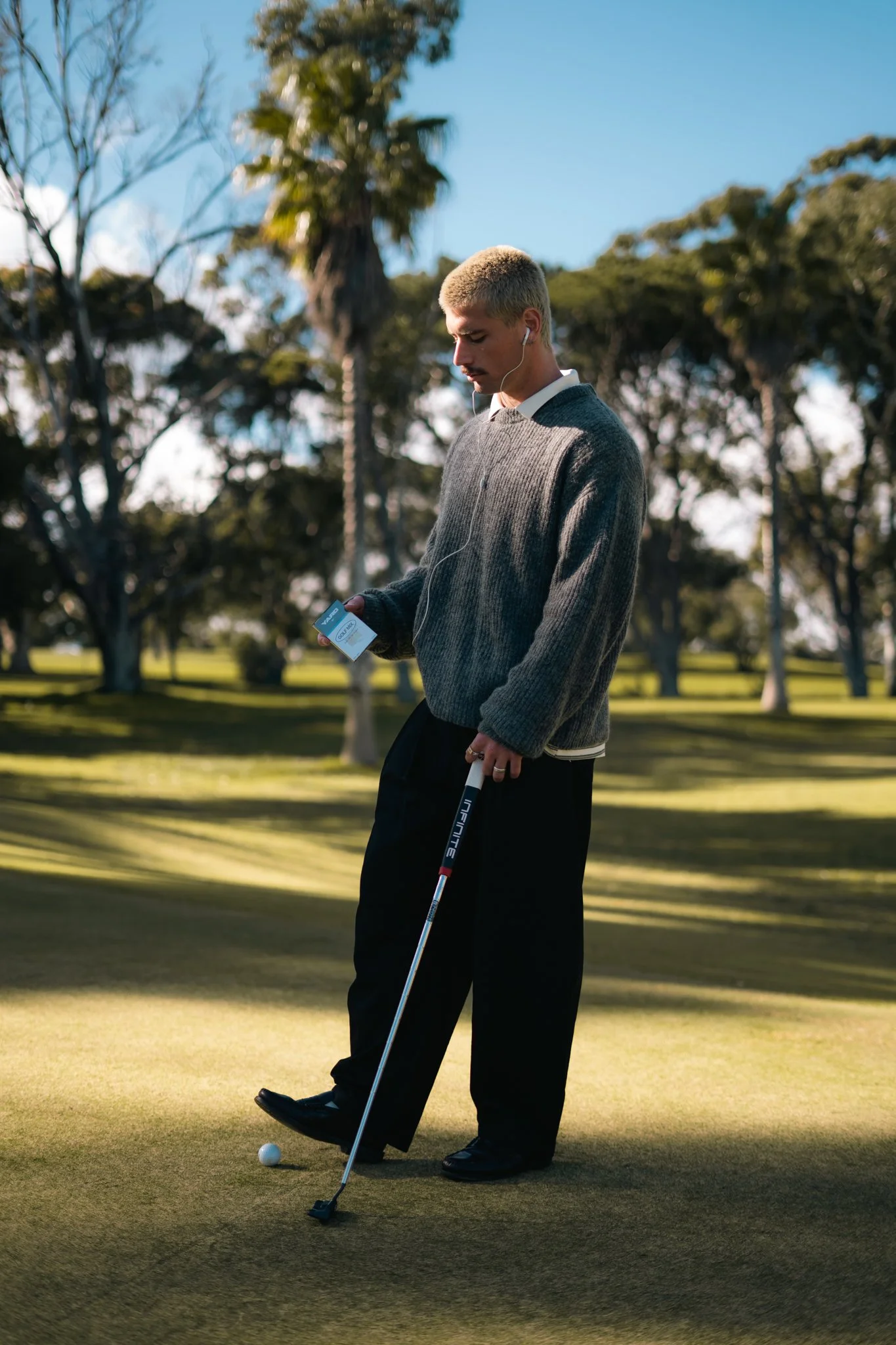 A man with blonde hair, dressed in a gray sweater and black pants, is playing golf on a green course. He is standing near the hole, holding a golf club, and looking down at the golf ball. He is wearing earphones and holding a small item in his left hand. The background features trees and a bright, clear sky.
