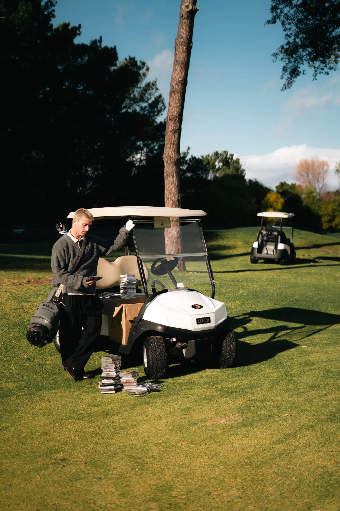 A young man with short blond hair dressed in a gray sweater and black pants standing next to a white golf cart on a golf course. The man is holding a golf tee and looking down at a stack of golf ball markers on the ground. There is a golf bag with clubs on the cart, and a pile of golf balls on the ground. A second golf cart is parked in the background near a large tree, and trees with green, yellow, and orange leaves are visible in the distance under a partly cloudy sky.