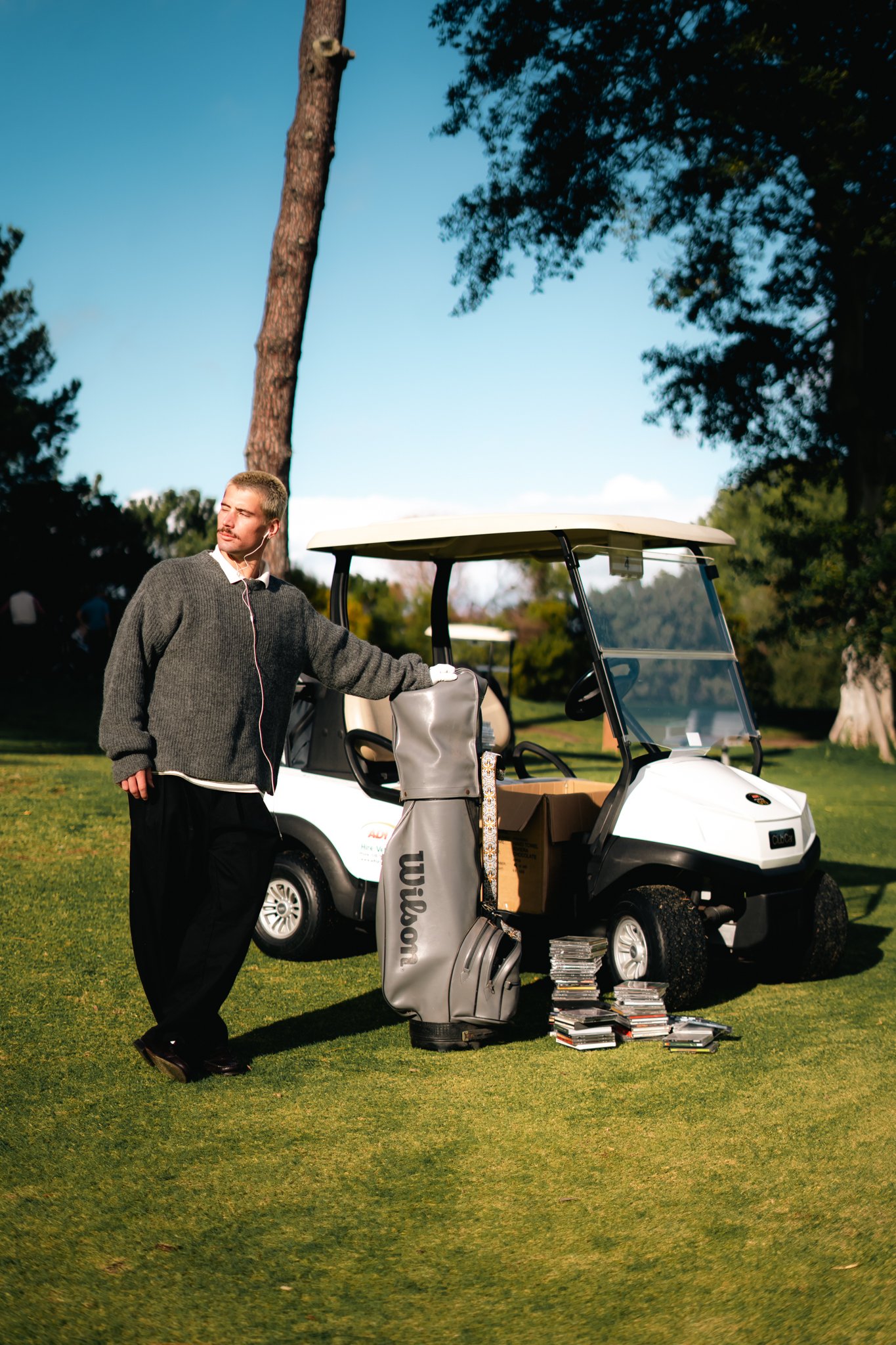 A man with blonde hair, wearing a grey sweater and black pants, stands next to a white golf cart on a golf course. He is holding and leaning on a golf bag filled with clubs, with a large pile of DVDs or CDs stacked on the ground nearby. The golf cart's windshield is visible, and the setting includes tall trees and a clear blue sky in the background.