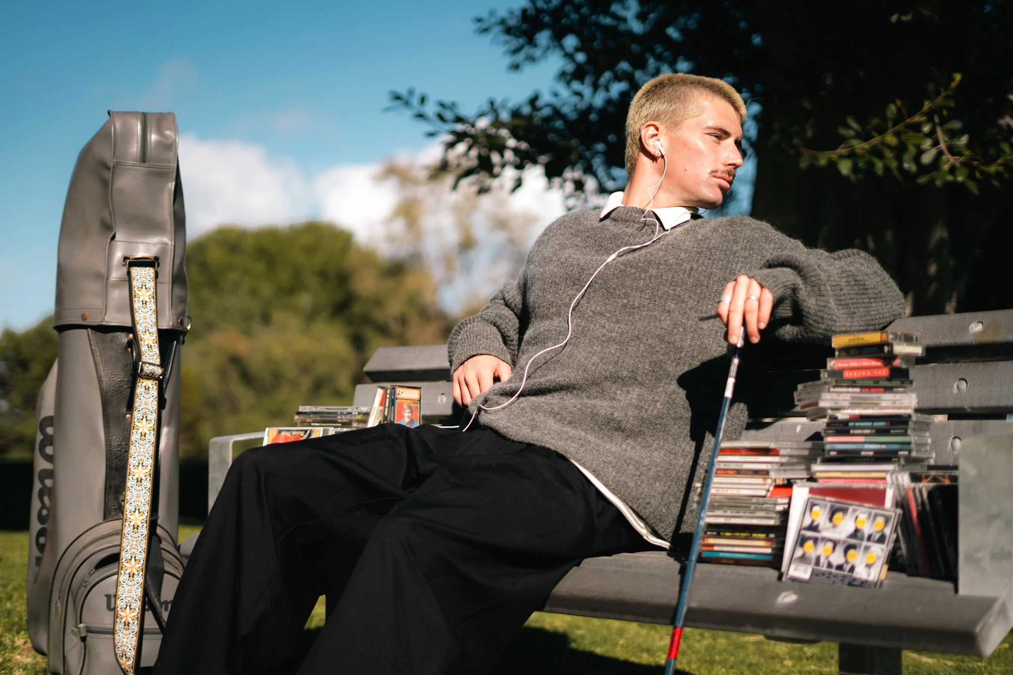 A man sitting on a park bench wearing a gray sweater, with short blond hair, and listening to music with white earbuds. A golf bag with a decorative strap and a stack of DVDs or CDs are beside him. He appears relaxed, looking away from the camera, with lush green trees and a blue sky in the background.