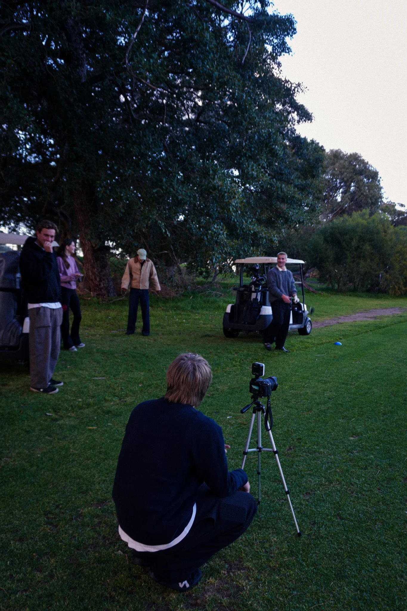 Group of people on a golf course, with some standing and one crouching near a camera on a tripod, and a man in a golf cart holding a golf club, with trees and grass in the background.