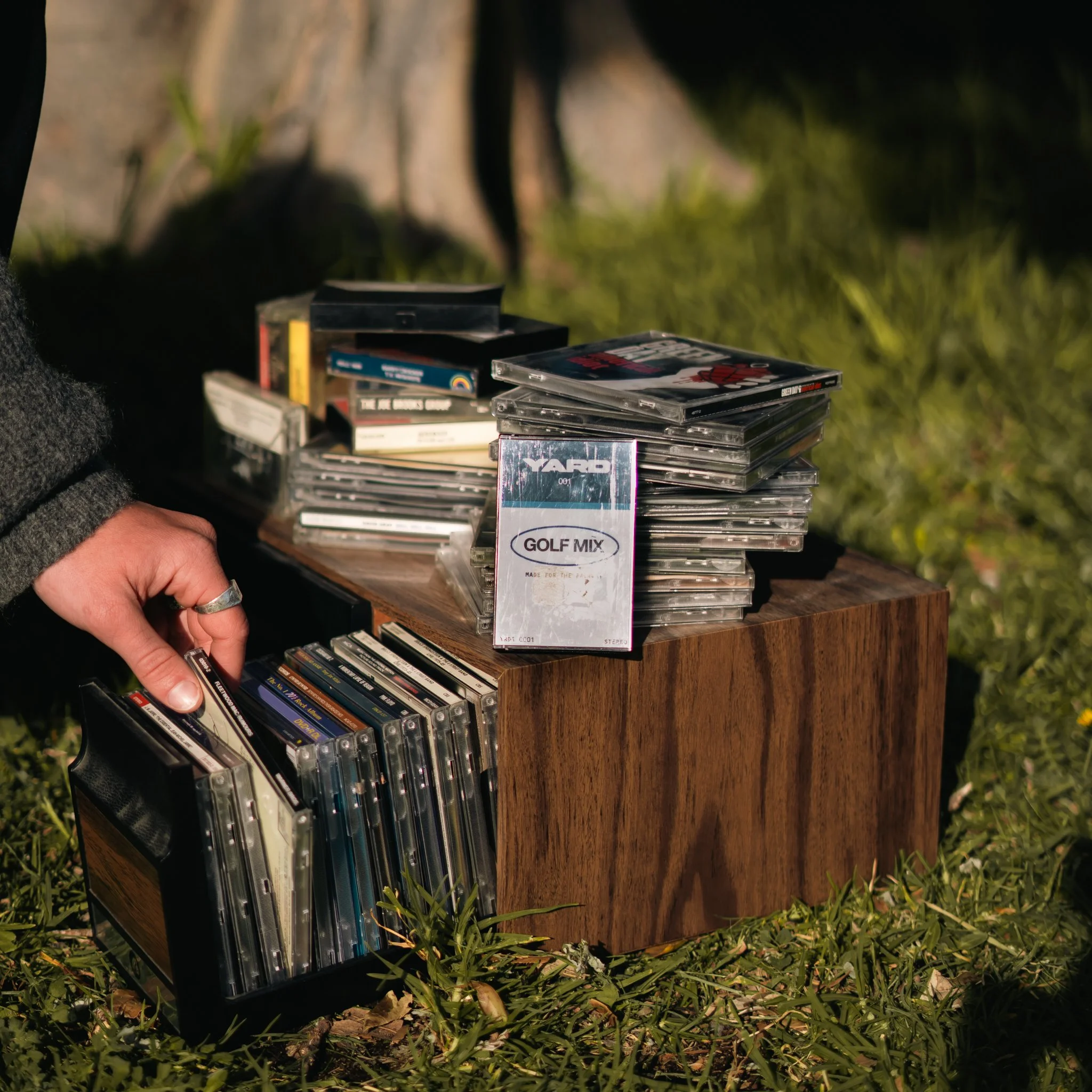 A stack of cassette tapes and some books on a wooden surface outdoors, with grass and plants around.