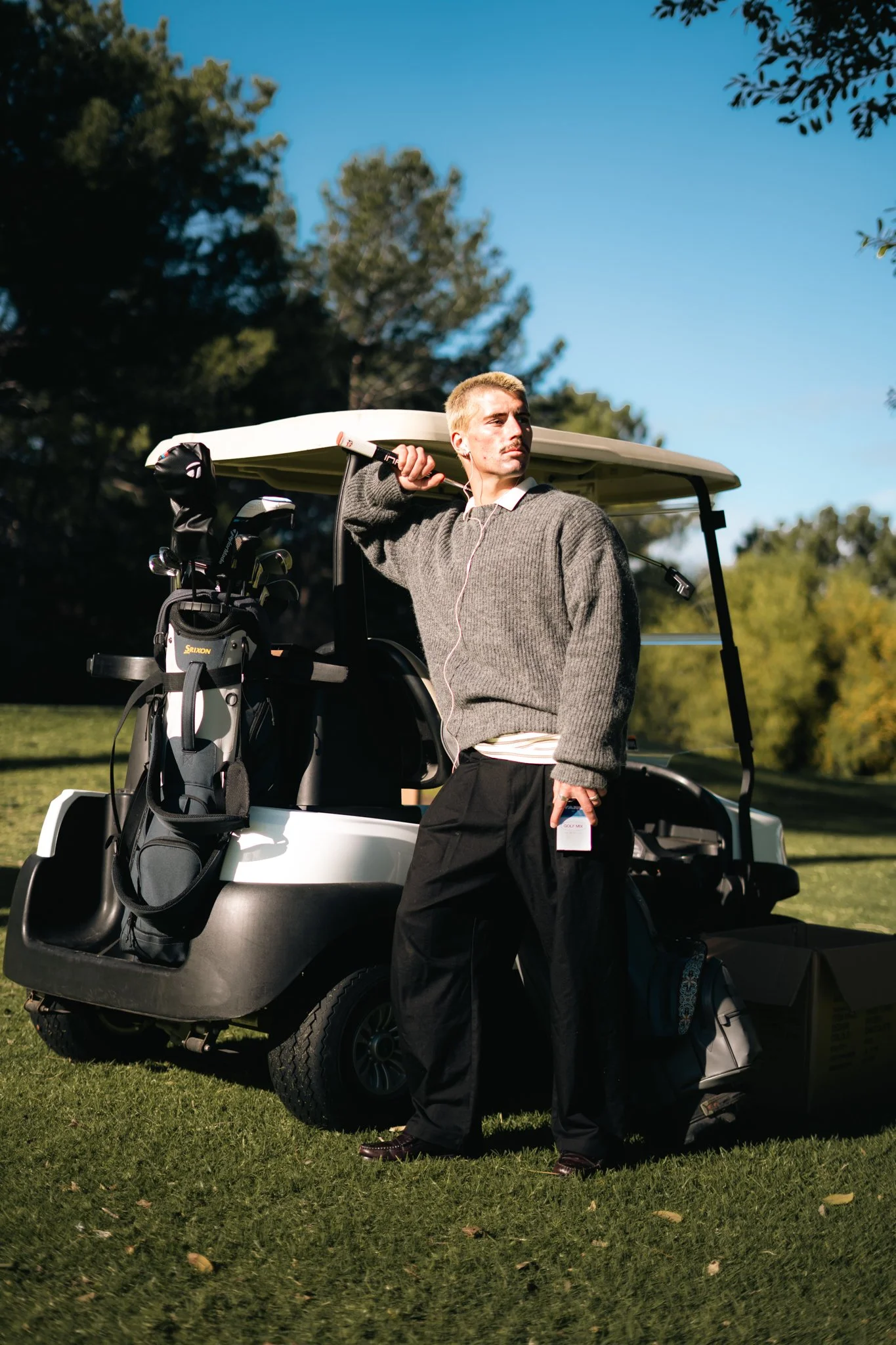 A man standing next to a golf cart on a golf course, holding a golf club and carrying a golf bag, with trees and a clear blue sky in the background.